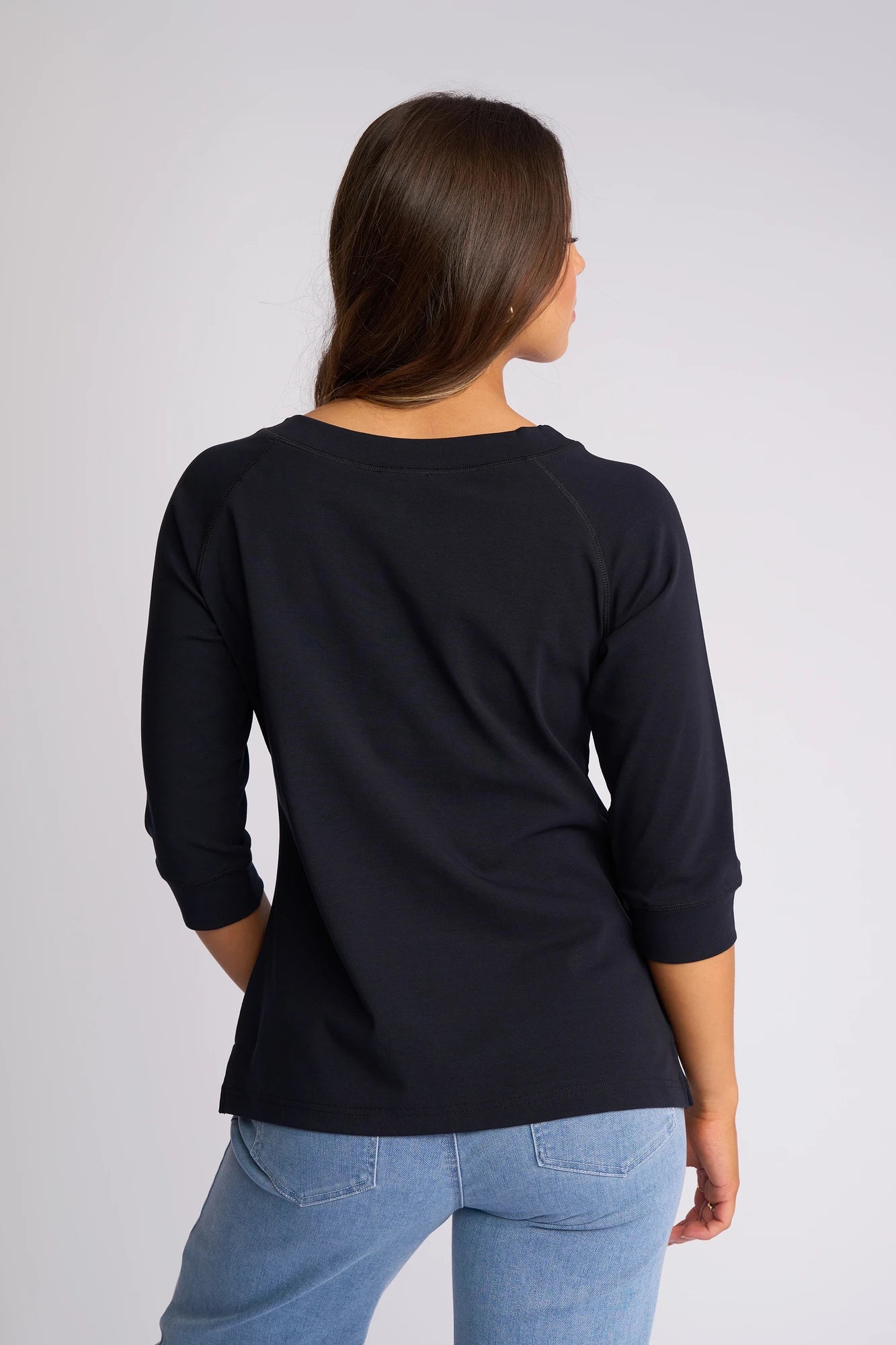 A person with long brown hair, wearing a Vassalli Canyon Top - Ink with 3/4 length sleeves and light blue jeans, is standing with their back to the camera against a plain background.