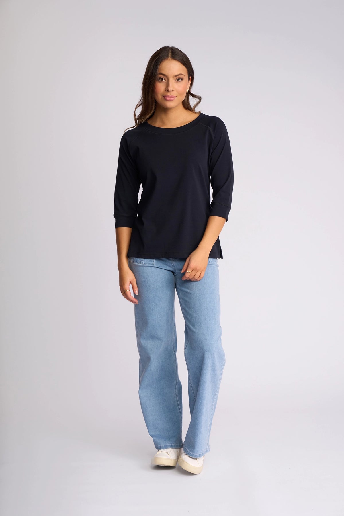 A woman stands against a simple light background, wearing the Vassalli Canyon Top - Ink, featuring 3/4 length sleeves and crafted from a comfortable cotton-elastane blend. She pairs it with light blue jeans and white shoes, her posture relaxed and her expression gentle.