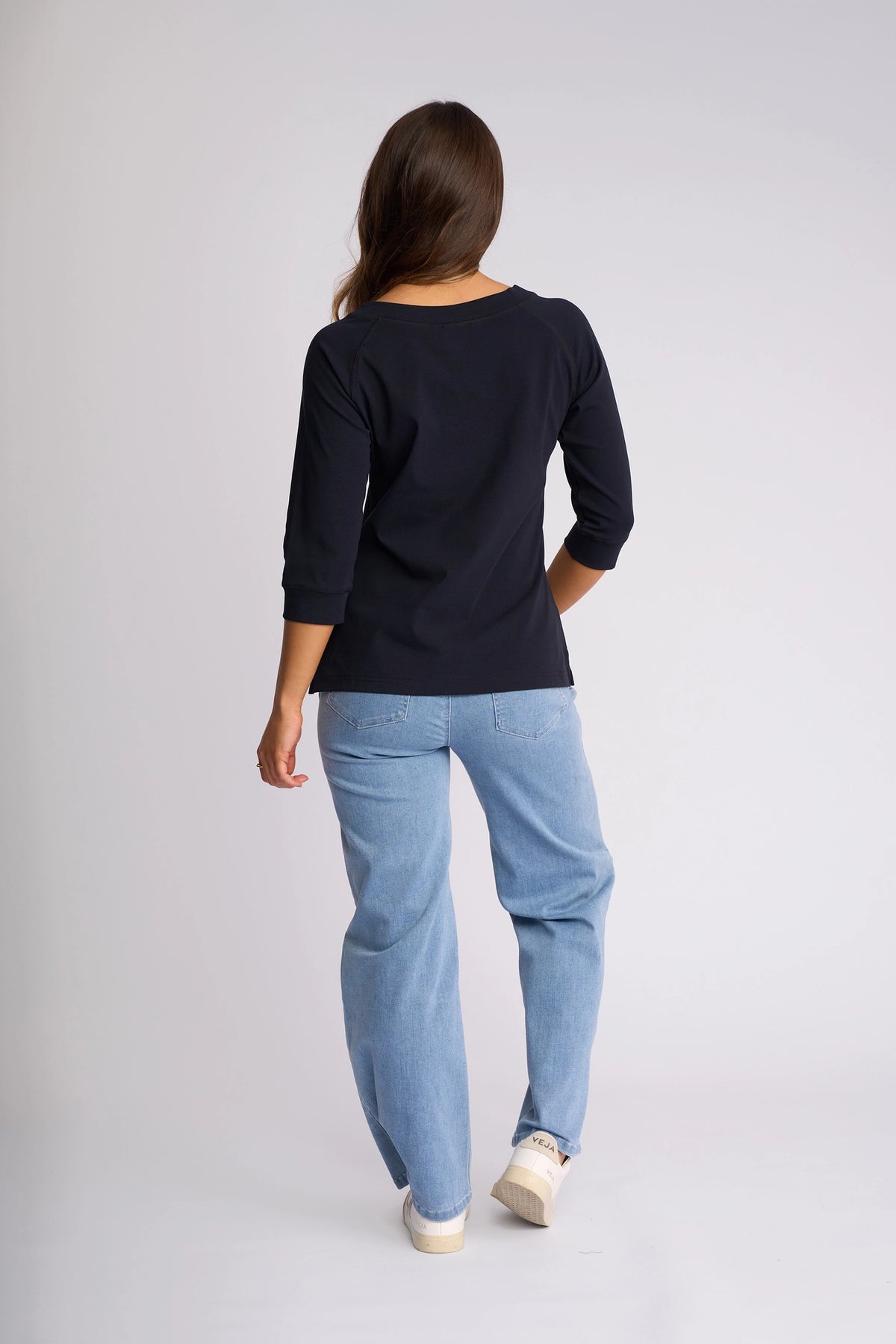 A person with long brown hair, wearing a black Vassalli Canyon Top - Ink with 3/4 length sleeves, light blue jeans, and white sneakers, stands facing away from the camera against a plain light background.