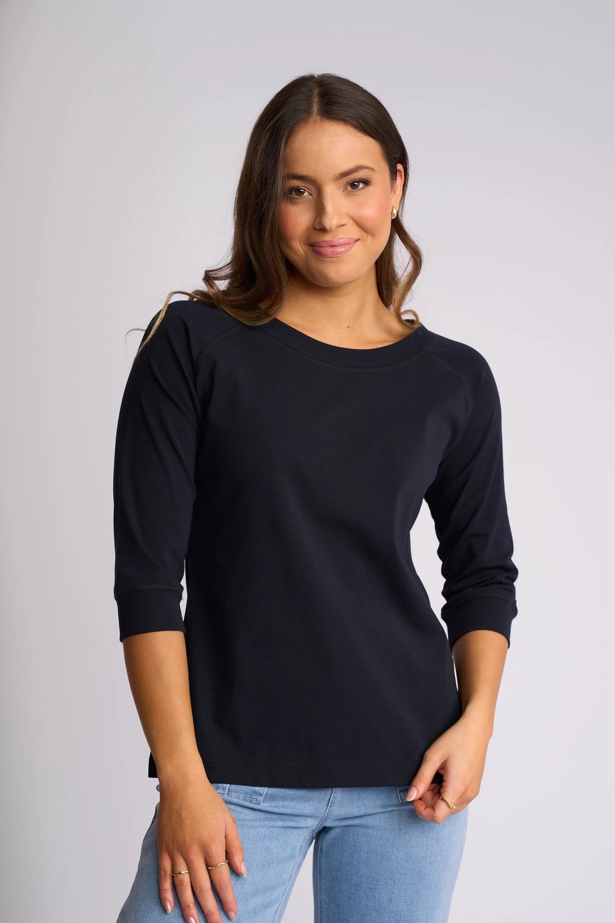A woman with long brown hair wearing the Vassalli Canyon Top in Ink, made from a comfortable cotton-elastane blend, paired with light blue jeans, stands against a plain light background and smiles softly at the camera.