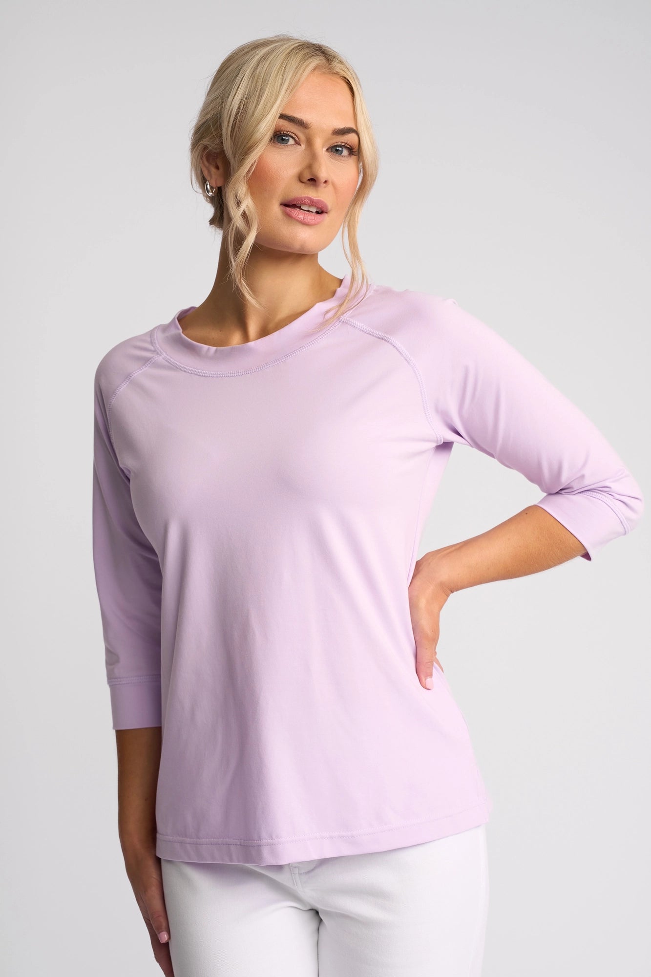 A woman with blonde hair styled in a low bun, wearing the Vassalli Canyon Top in light purple—crafted from a comfortable cotton-elastane blend—and white pants, stands against a plain light background with one hand on her hip, smiling slightly.