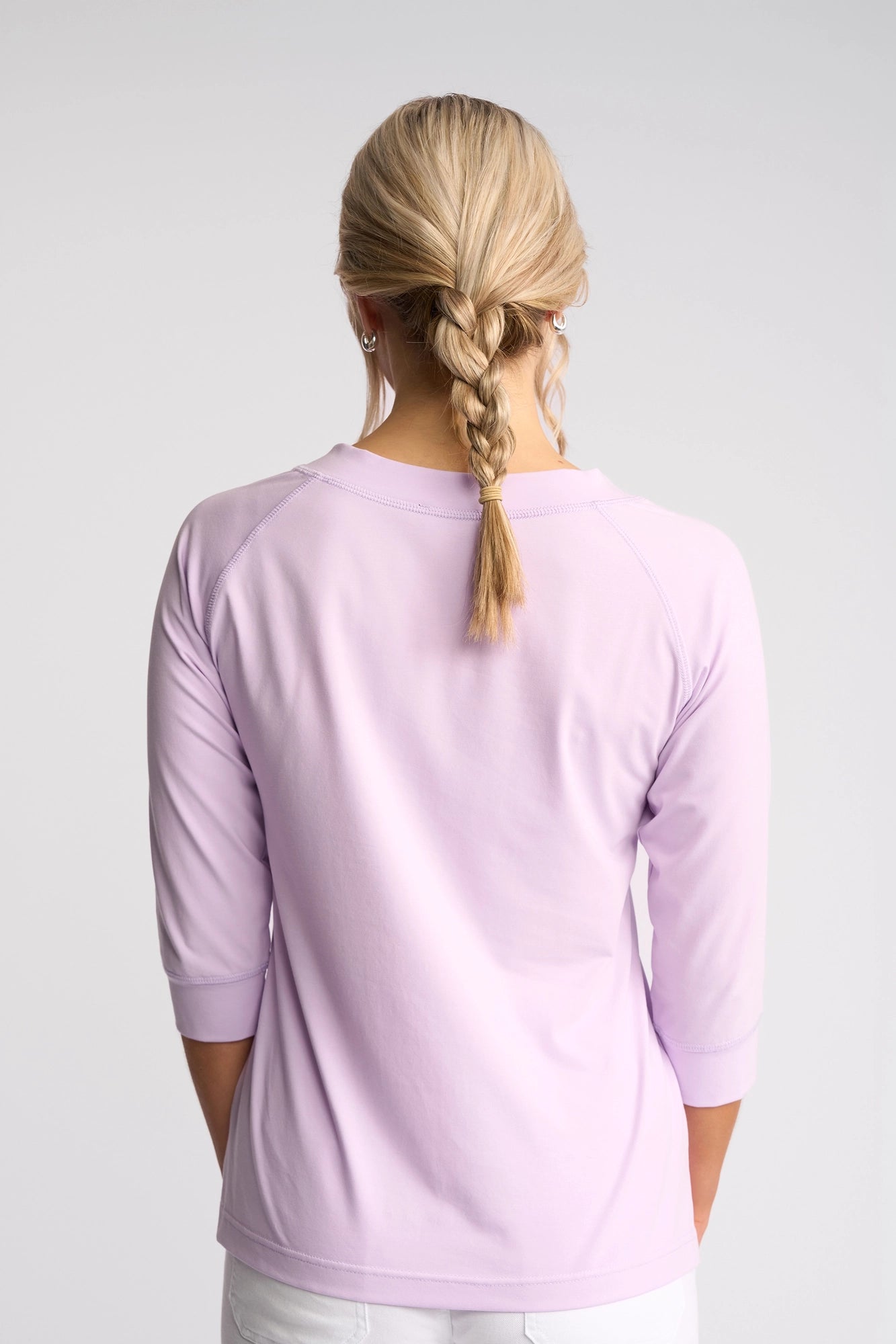 A person with blonde braided hair, dressed in Vassalli’s Canyon Top in Light Purple—a comfortable cotton-elastane blend—and white pants, is shown from the back against a plain light background.