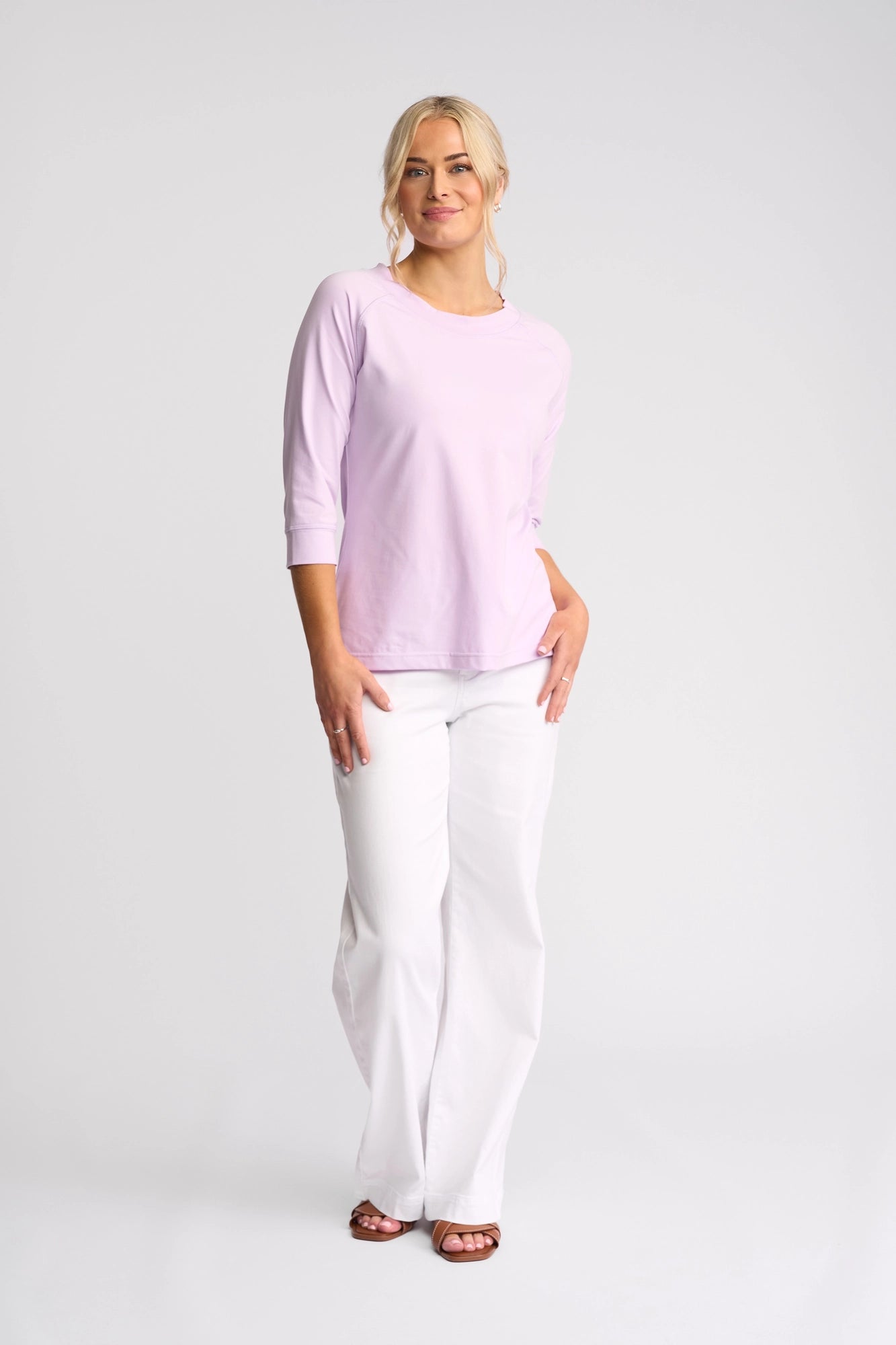 A woman stands smiling, wearing the Vassalli Canyon Top - Light Purple with three-quarter sleeves, paired with white pants and brown sandals, set against a plain light background.