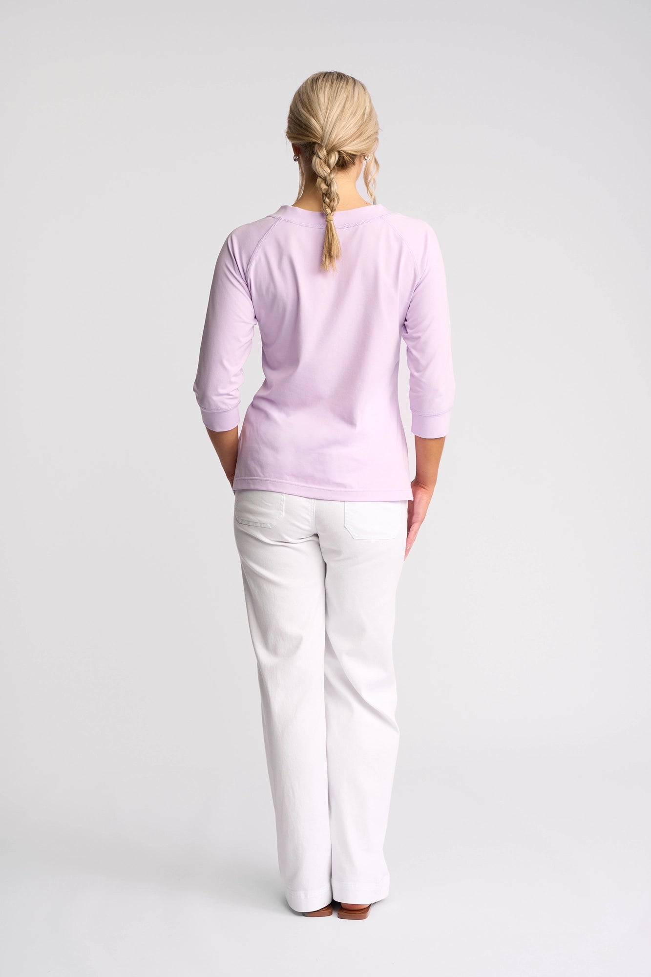 A person with blonde, braided hair wearing the Vassalli Canyon Top - Light Purple, made from a soft cotton-elastane blend, paired with white pants, stands facing away from the camera against a plain white background.
