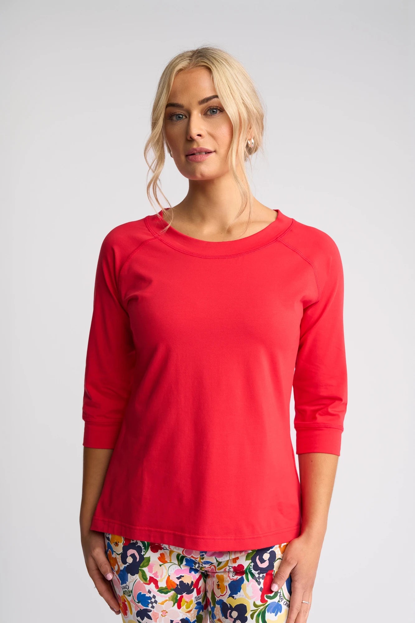 A woman with blonde hair wears a Vassalli Canyon Top in Raspberry, featuring 3/4 length sleeves, paired with colorful floral pants. She stands against a plain light background, showcasing the top’s comfortable and stylish cotton-elastane blend.