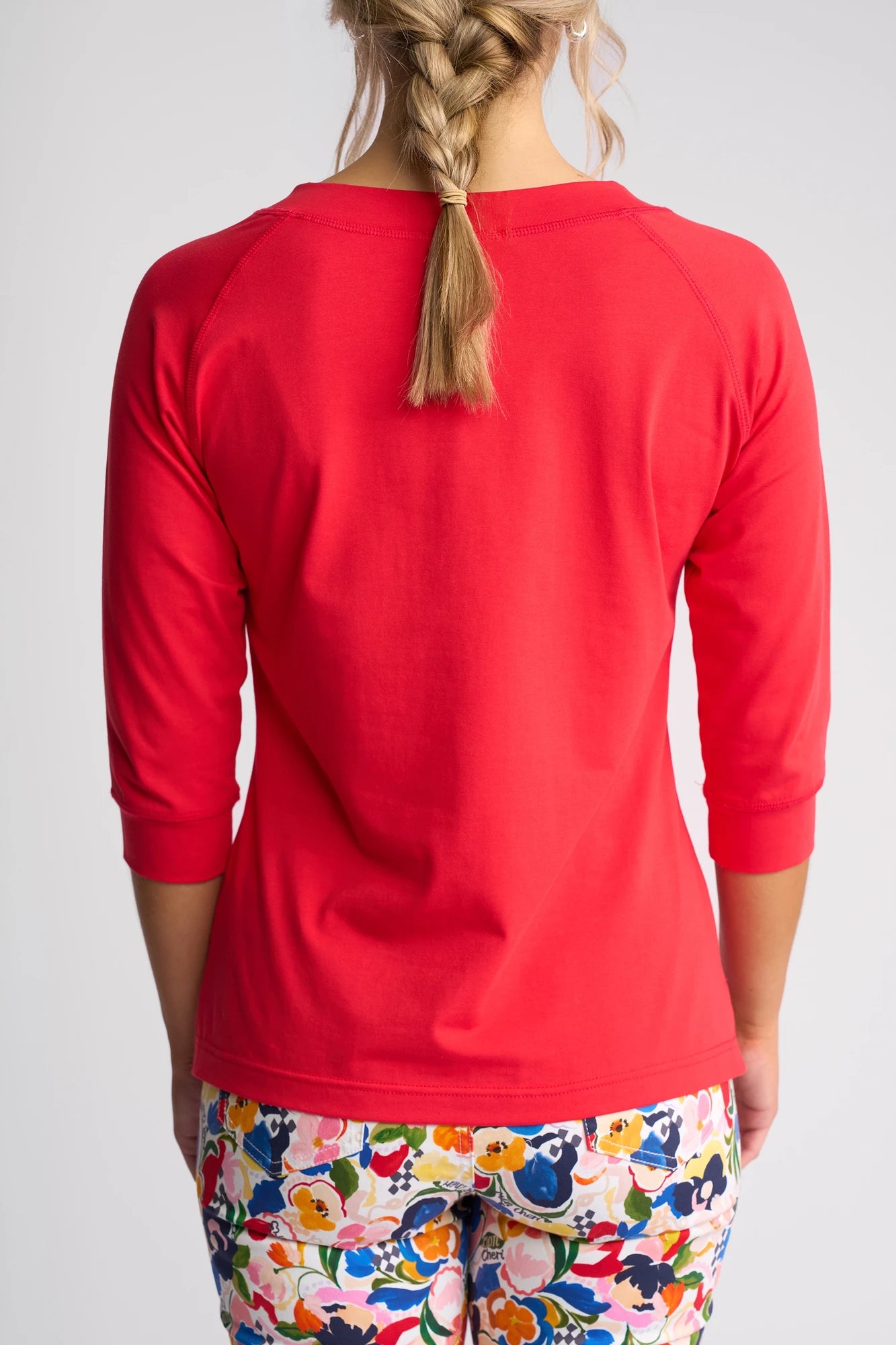 A person with a braid is shown from the back, wearing the Vassalli Canyon Top - Raspberry with 3/4 length sleeves, paired with pants featuring a colorful floral pattern in a comfortable cotton-elastane blend.