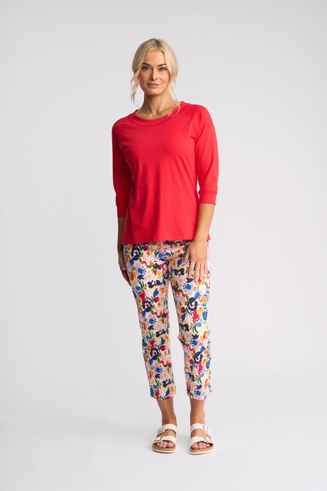 A woman stands against a plain background, wearing a bright raspberry Vassalli Canyon Top with 3/4 length sleeves, paired with colorful floral patterned pants and white sandals. Her blond hair is tied back and she is smiling slightly.