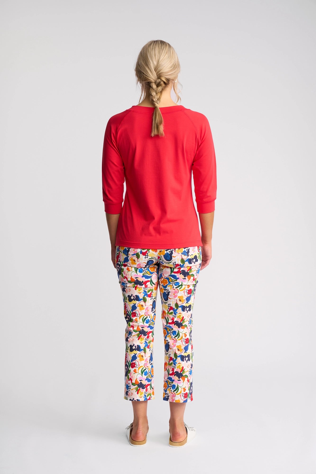 A woman stands facing away, wearing the Vassalli Canyon Top in Raspberry with 3/4 length sleeves, paired with colorful, floral-patterned pants. Her blonde hair is styled in a loose braid and she is barefoot against a plain white background. Crafted from a comfortable cotton-elastane blend, the top provides an easy fit and stylish look.