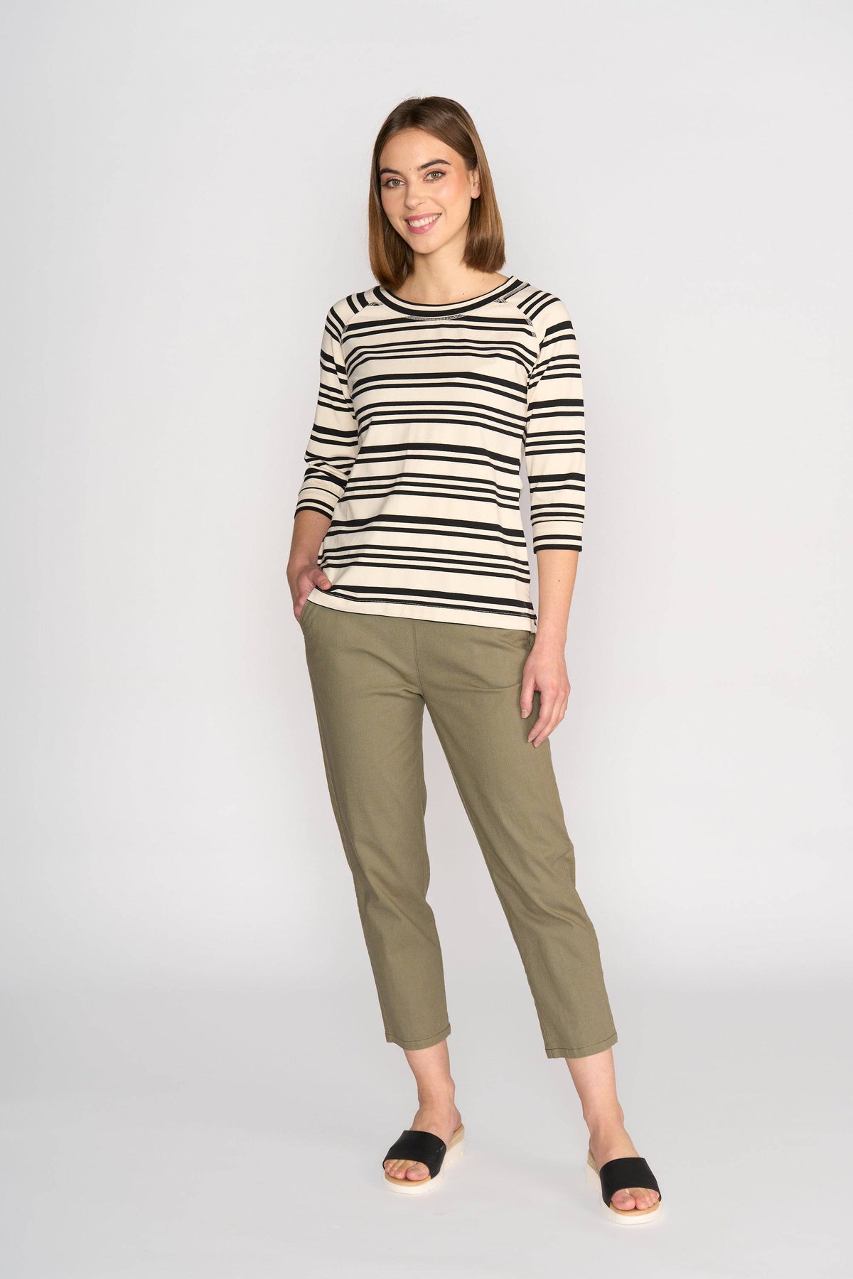 A woman smiles against a light background, wearing the Vassalli Canyon Top in Ecru/Black Stripe with olive green pants and black slides. One hand is in her pocket, the other by her side.