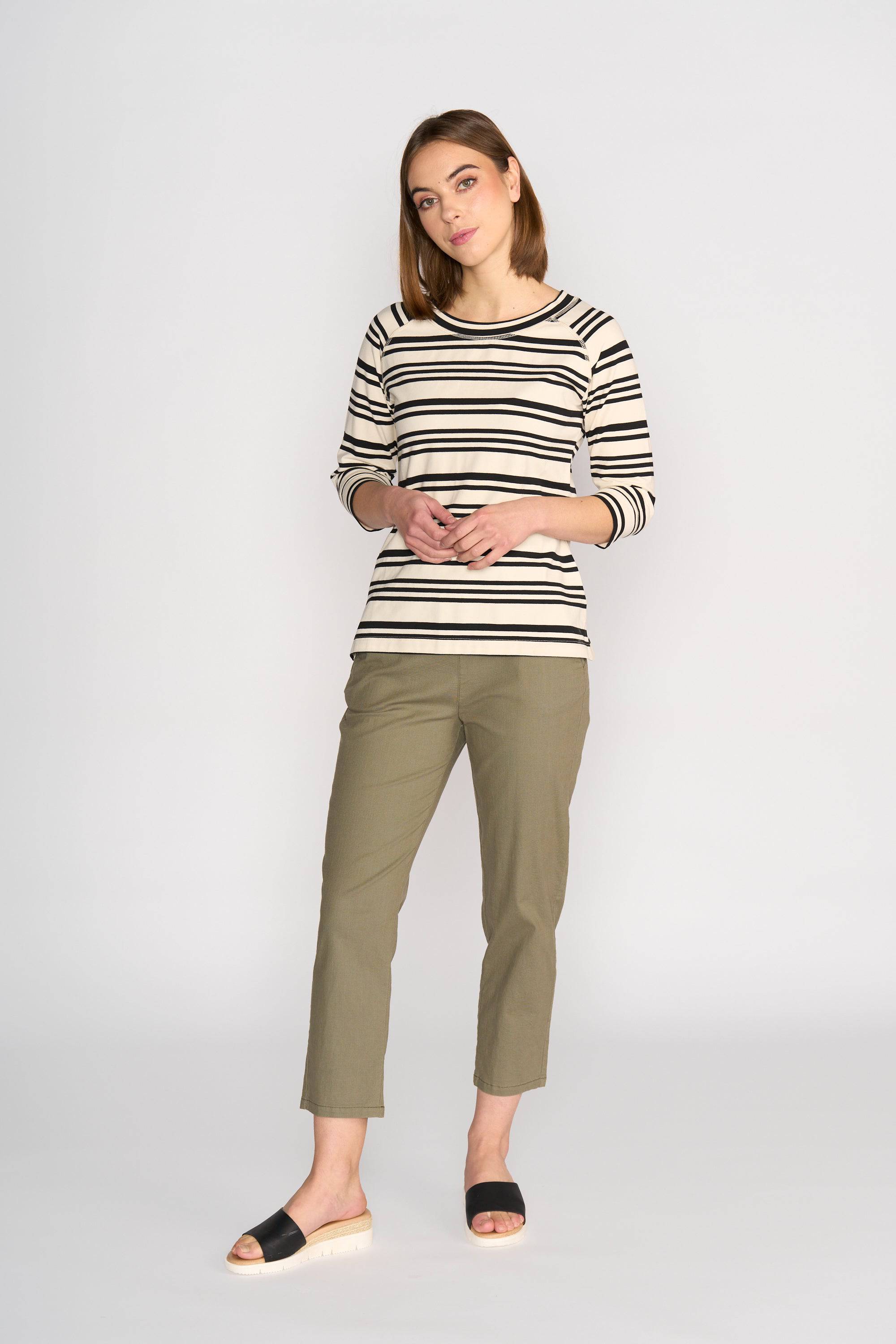A woman poses casually against a plain background wearing the Vassalli Canyon Top in Ecru/Black Stripe with olive green cropped pants and black slide sandals. She has straight, shoulder-length hair and a relaxed style.