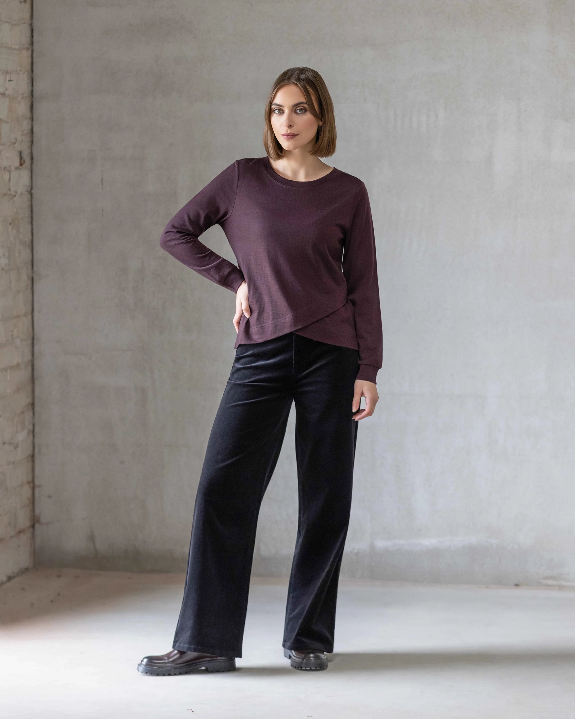 A woman with short brown hair stands indoors against a light gray concrete wall, wearing the Vassalli Remy Merino Top in Berry with a layered crossover hem, loose black pants, and black shoes. She poses neutrally with one hand on her hip.