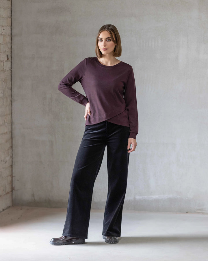 A woman with short brown hair stands indoors against a light gray concrete wall, wearing the Vassalli Remy Merino Top in Berry with a layered crossover hem, loose black pants, and black shoes. She poses neutrally with one hand on her hip.