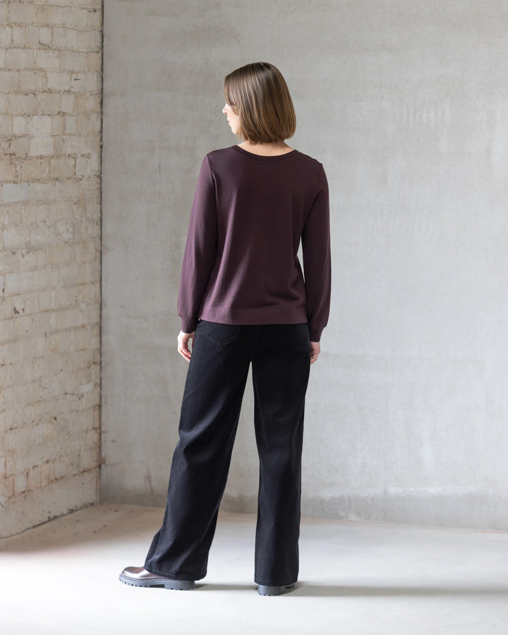 A woman with short brown hair stands indoors facing a concrete wall, wearing the Vassalli Remy Merino Top in Berry, loose black pants, and gray shoes. Bright light casts soft shadows on the floor and wall.