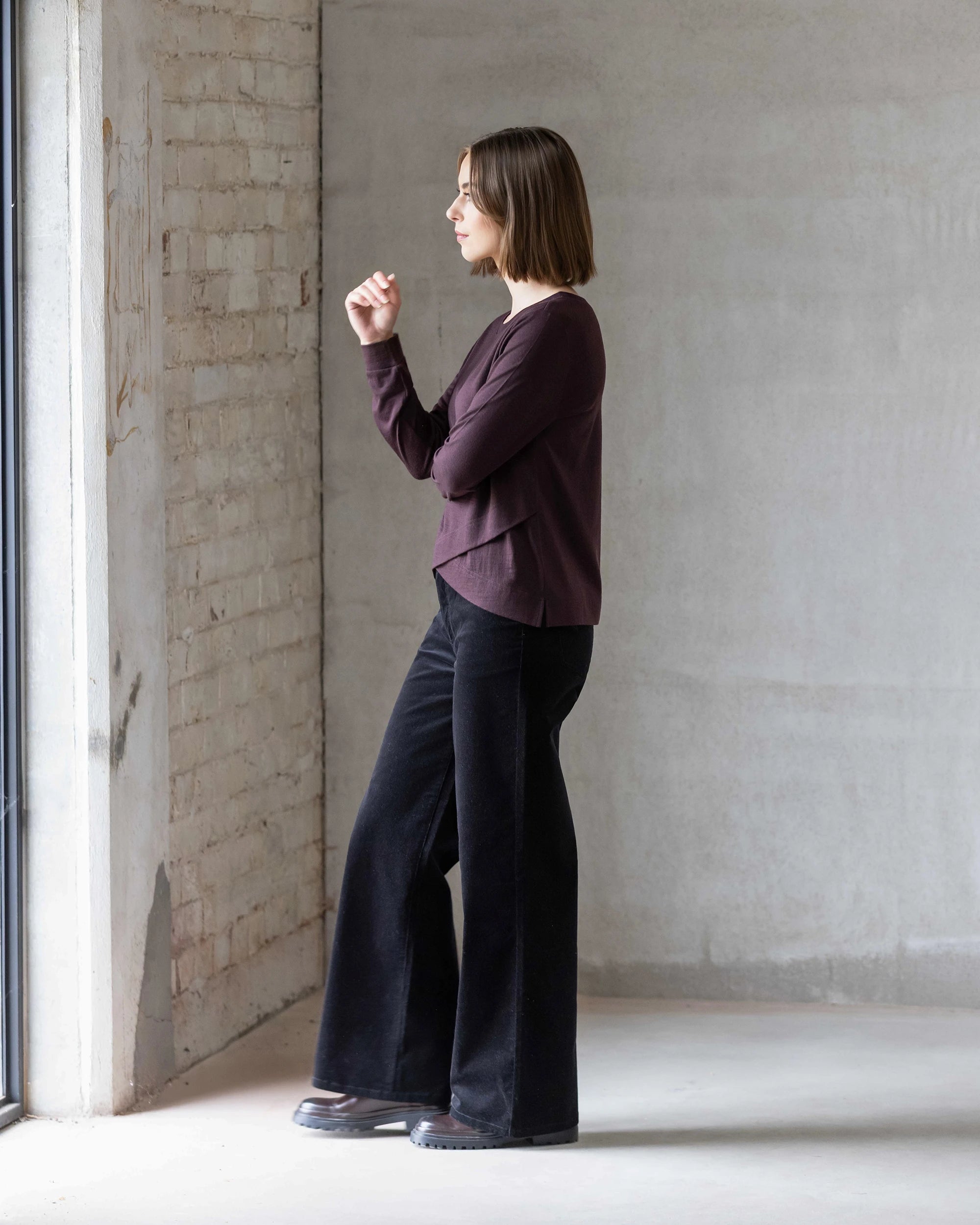 A woman with shoulder-length hair stands in a minimalist room, wearing the Vassalli Remy Merino Top in Berry, paired with black wide-leg pants and shoes, gazing out a window. Perfect for everyday wear.