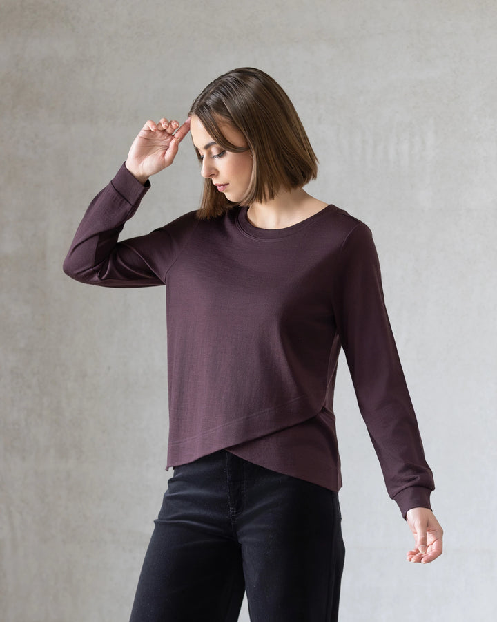 A woman with chin-length brown hair wears the Vassalli Remy Merino Top in Berry and black pants, standing before a light gray background with one arm raised, capturing effortless everyday style.