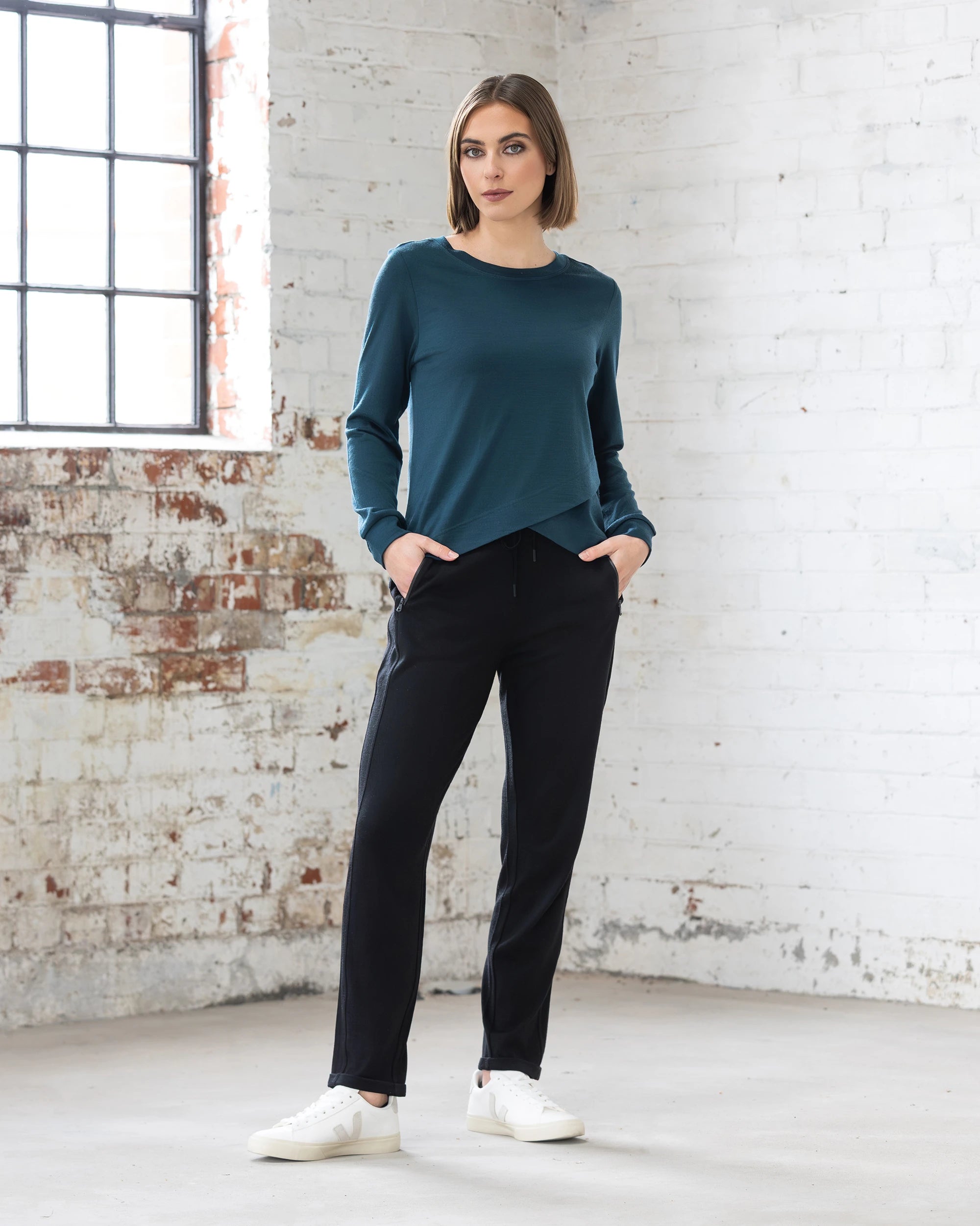 A woman stands in a bright room with white brick walls, wearing the Vassalli Remy Merino Top in Dragonfly, black pants, and white sneakers. She has her hands in her pockets and looks confidently at the camera.