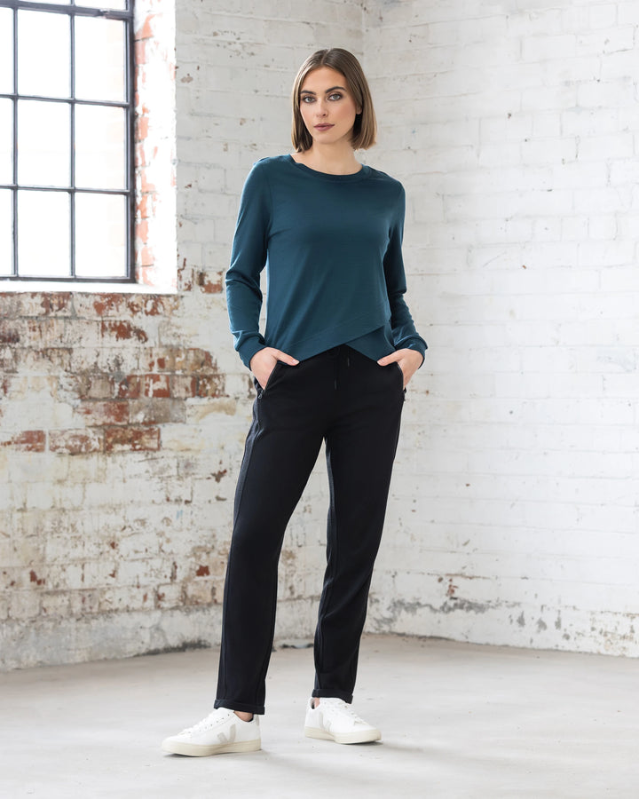 A woman stands in a bright room with white brick walls, wearing the Vassalli Remy Merino Top in Dragonfly, black pants, and white sneakers. She has her hands in her pockets and looks confidently at the camera.