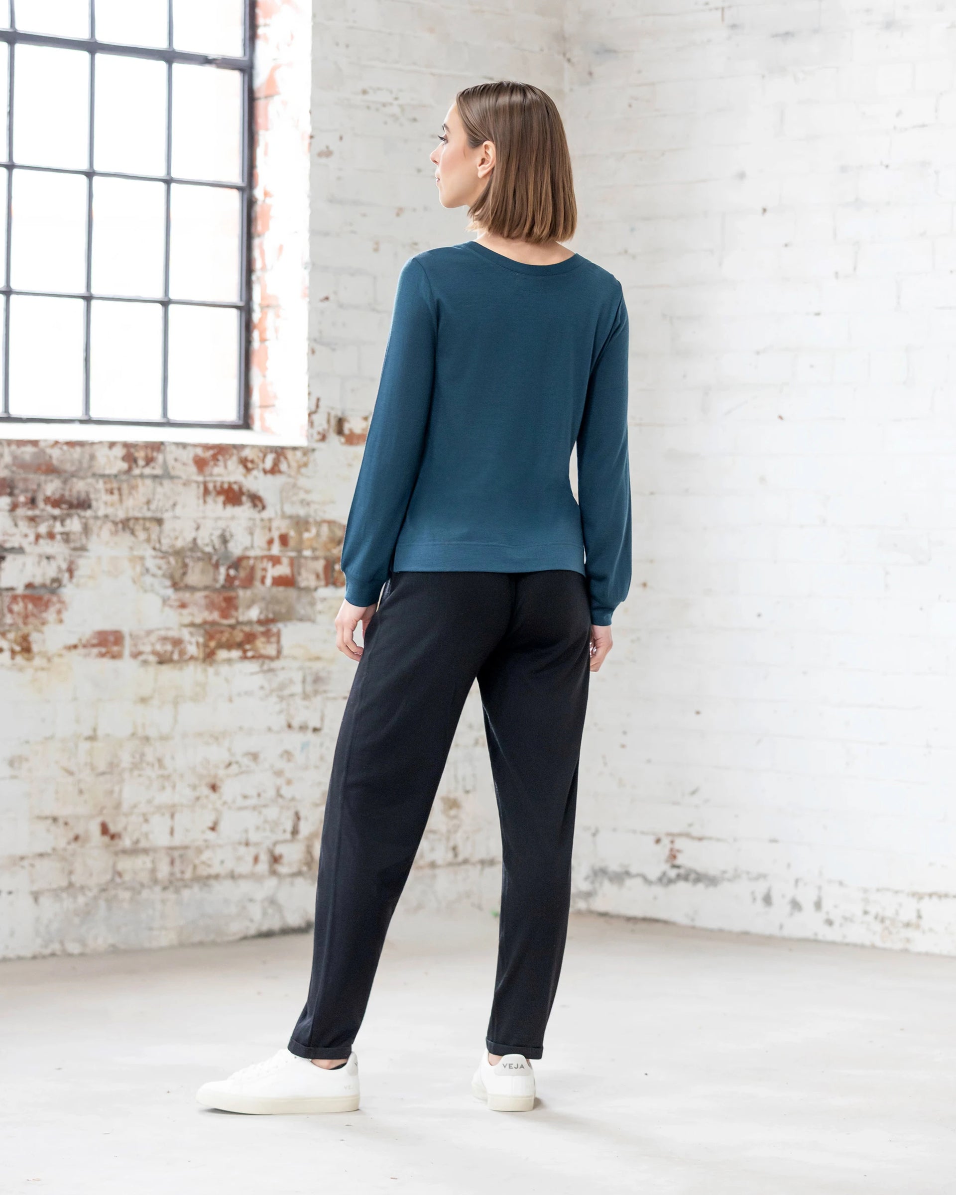 A person with straight, shoulder-length hair stands indoors facing away, wearing the Vassalli Remy Merino Top - Dragonfly—a cozy merino long-sleeve in dark teal—paired with black pants and white sneakers, perfect for winter layering.