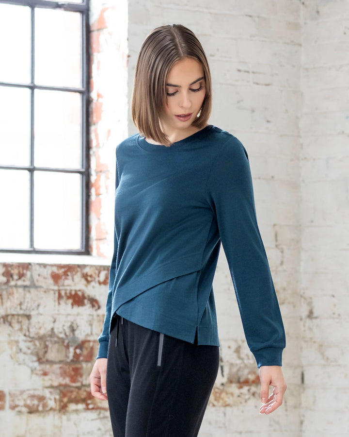 A woman with straight, shoulder-length brown hair wears the Vassalli Remy Merino Top in Dragonfly and black pants. She stands indoors by a large window, looking down against white-painted brick walls.