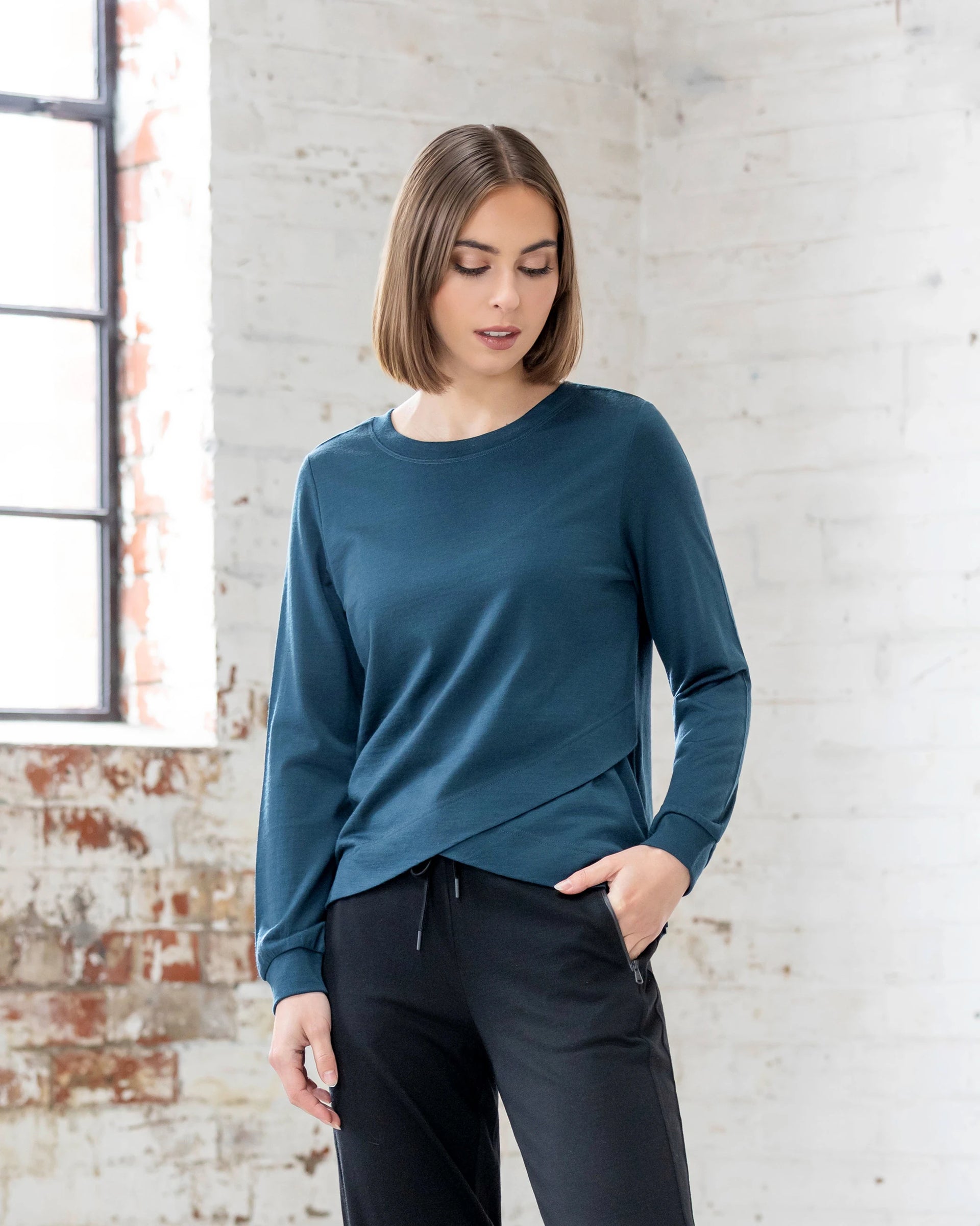 A woman with a bob haircut wears the Vassalli Remy Merino Top - Dragonfly and black pants indoors, standing near a white brick wall and large window with one hand in her pocket—a perfect winter layer.