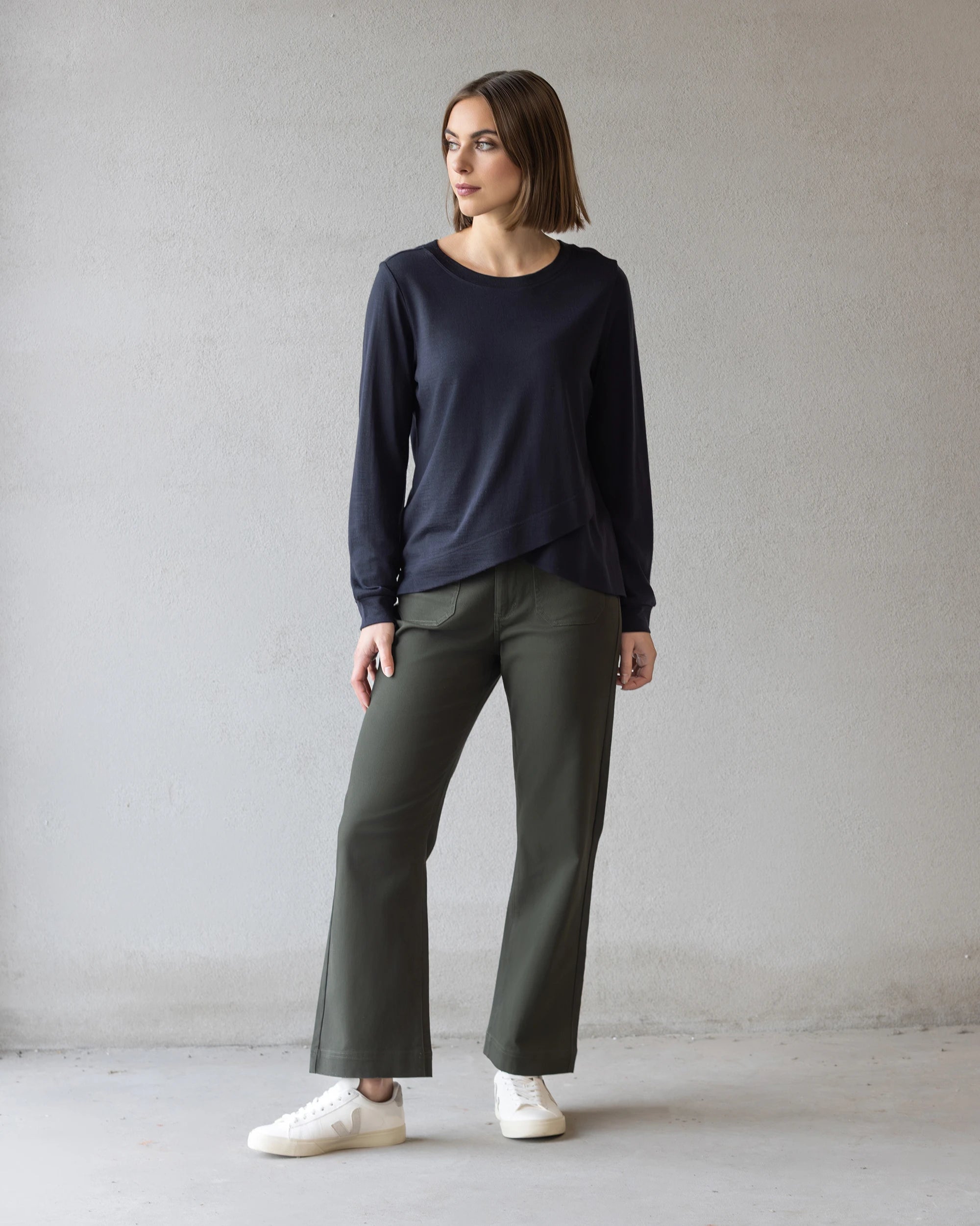 A woman with a straight brown bob wears the Vassalli Remy Merino Top in Midnight, paired with olive wide-leg pants and white sneakers, standing against a plain light gray wall and looking to her left.
