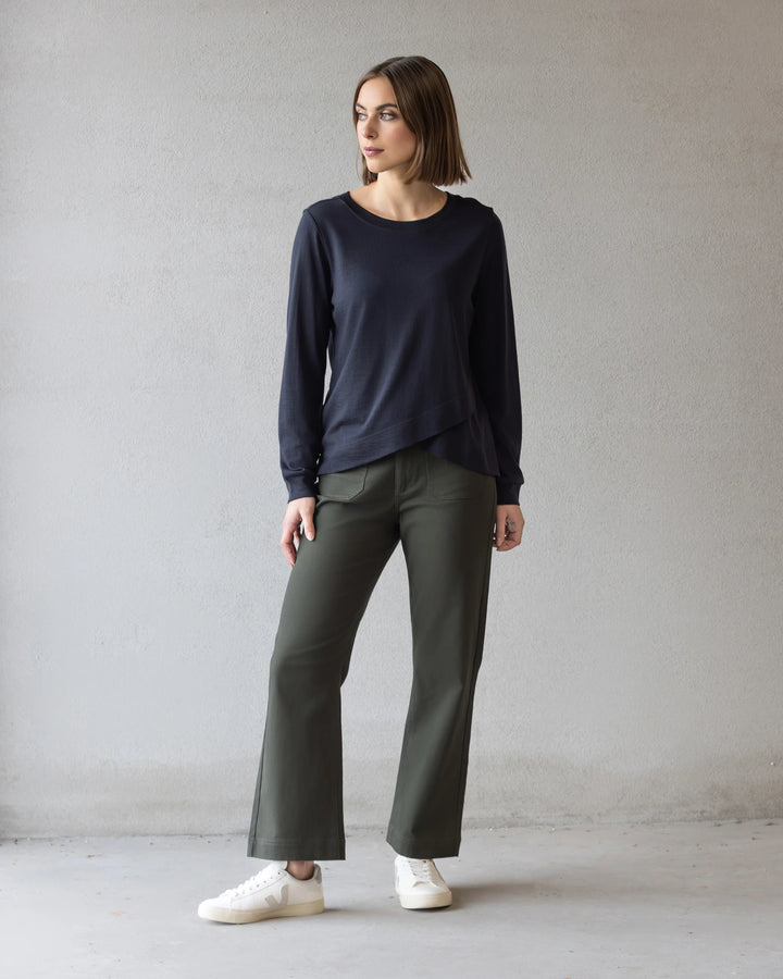 A woman with a straight brown bob wears the Vassalli Remy Merino Top in Midnight, paired with olive wide-leg pants and white sneakers, standing against a plain light gray wall and looking to her left.