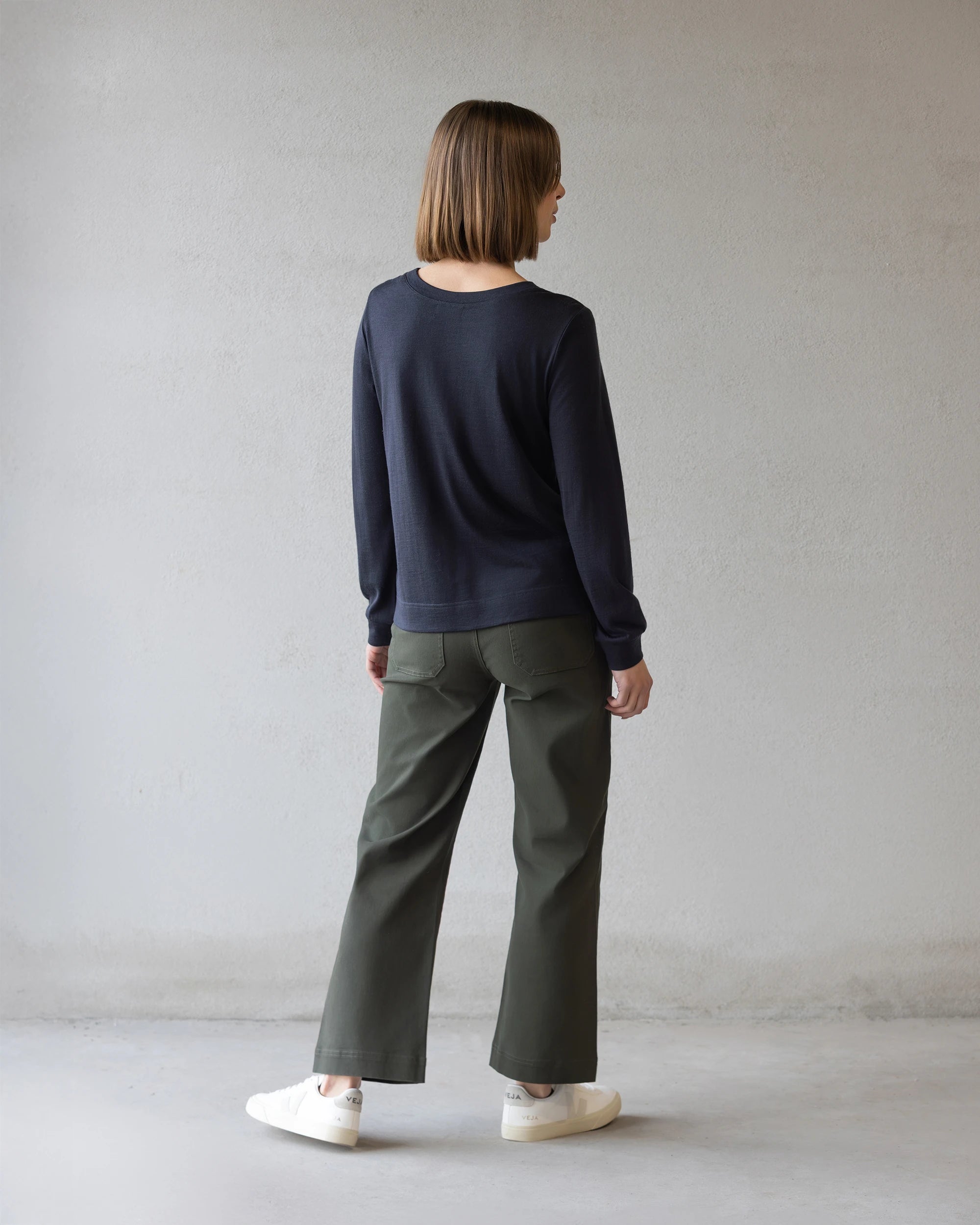 A person with straight, shoulder-length brown hair faces away wearing the Vassalli Remy Merino Top in Midnight, olive green wide-leg pants, and white sneakers against a plain light gray wall.