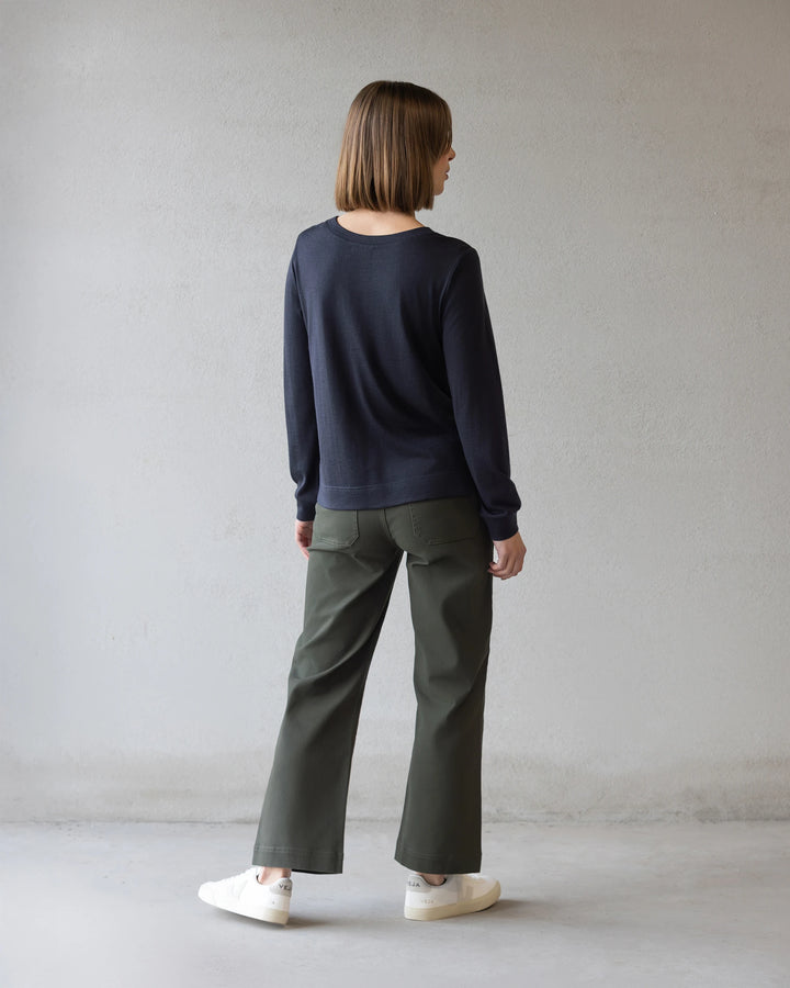 A person with straight, shoulder-length brown hair faces away wearing the Vassalli Remy Merino Top in Midnight, olive green wide-leg pants, and white sneakers against a plain light gray wall.