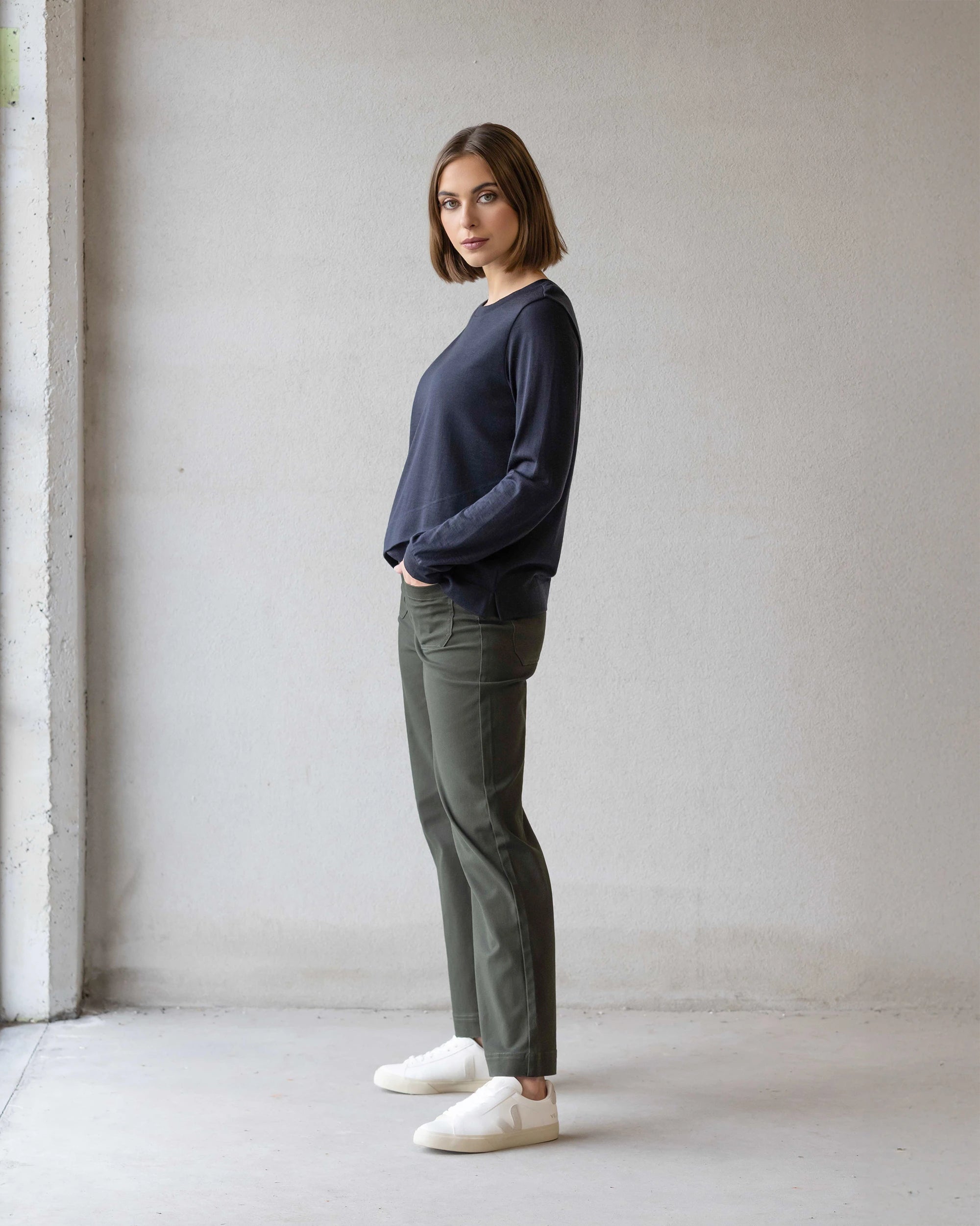 A woman with straight, shoulder-length brown hair stands sideways, wearing the Vassalli Remy Merino Top in Midnight, olive pants, and white sneakers in a minimalist indoor setting with a neutral-colored wall.