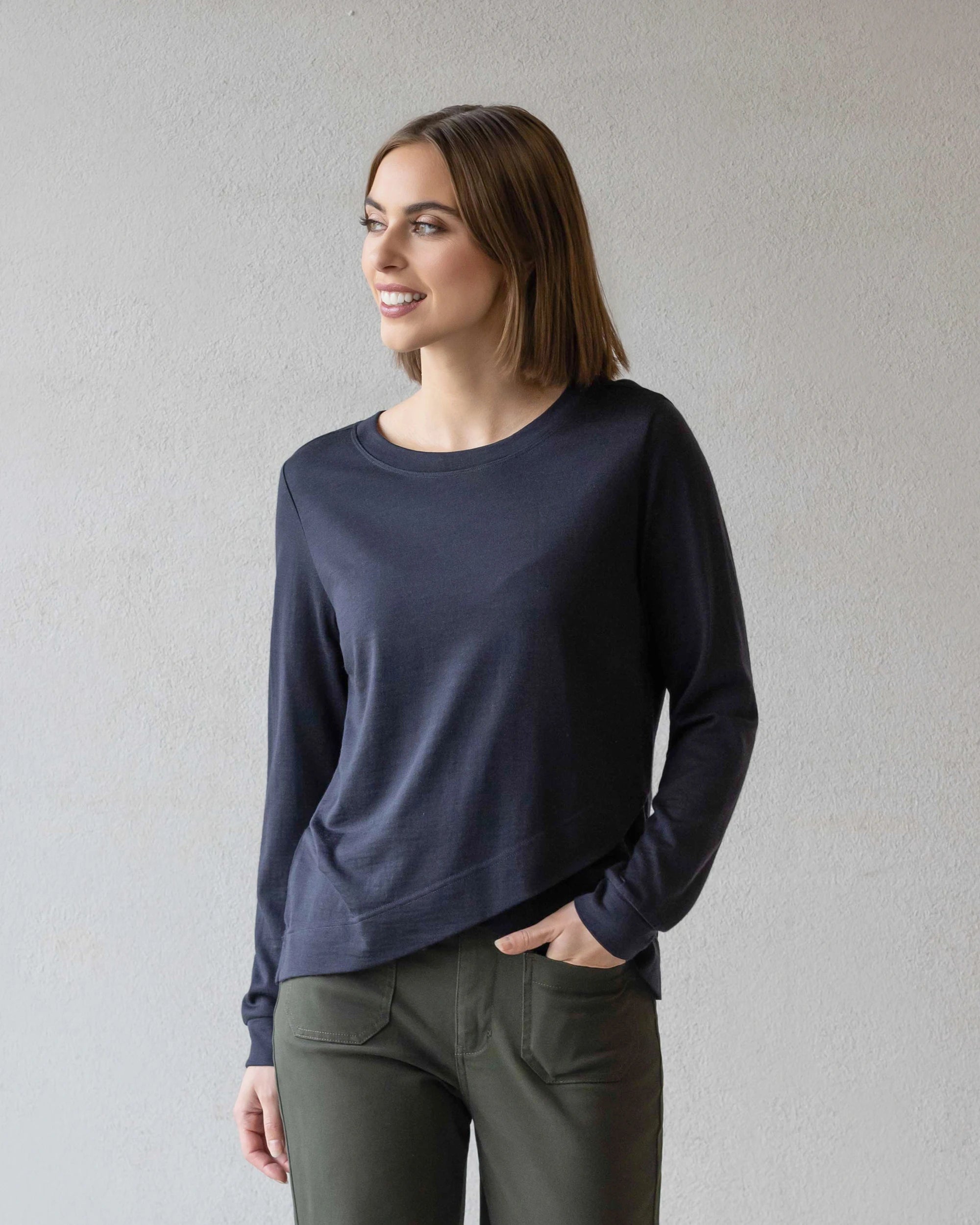 A woman with straight brown hair wears a Vassalli Remy Merino Top in Midnight and olive green pants, standing against a light gray wall. She smiles and looks to her left, one hand in her pocket.