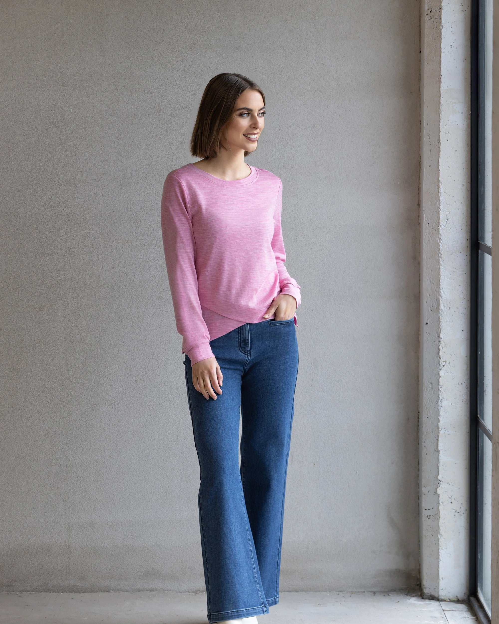A woman with a bob stands indoors near a window, smiling. She’s wearing the Vassalli Remy Merino Top in Pink Marle and blue jeans—ideal for winter layering—with one hand in her pocket against a plain gray wall.