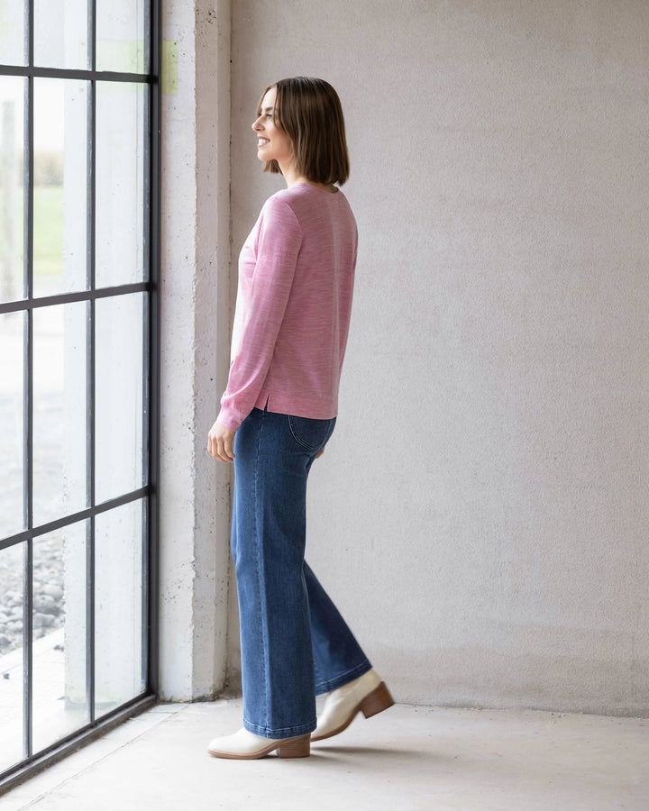 A woman wearing the Vassalli Remy Merino Top in Pink Marle, blue jeans, and beige shoes stands smiling by a large window, enjoying winter layering in a bright, minimalist room.
