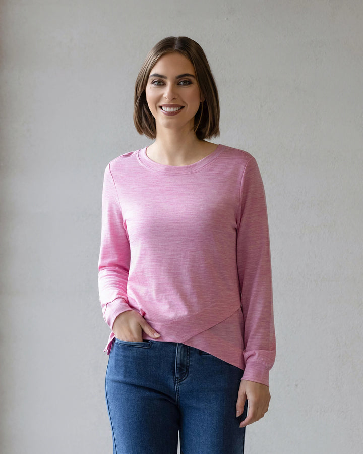 A woman with short brown hair smiles at the camera, wearing the Vassalli Remy Merino Top in Pink Marle and blue jeans, ideal for winter layering, while standing against a plain light gray background.