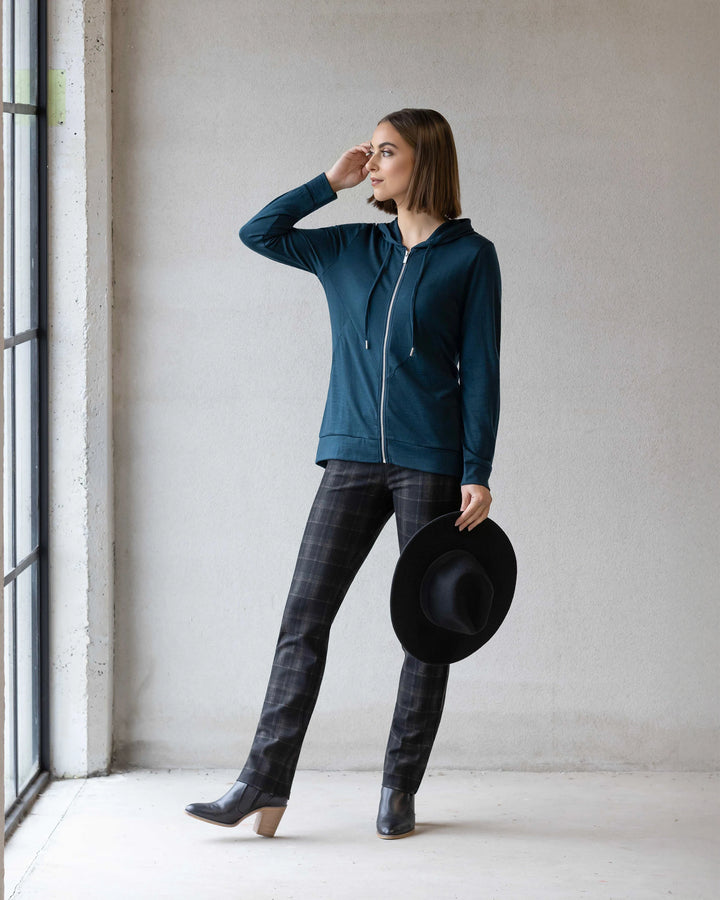 A woman in the Vassalli Shield Merino Hoodie in Dragonfly stands indoors, holding a black hat and gazing out a large window, with plaid pants, ankle boots, and a neutral-colored wall behind her.