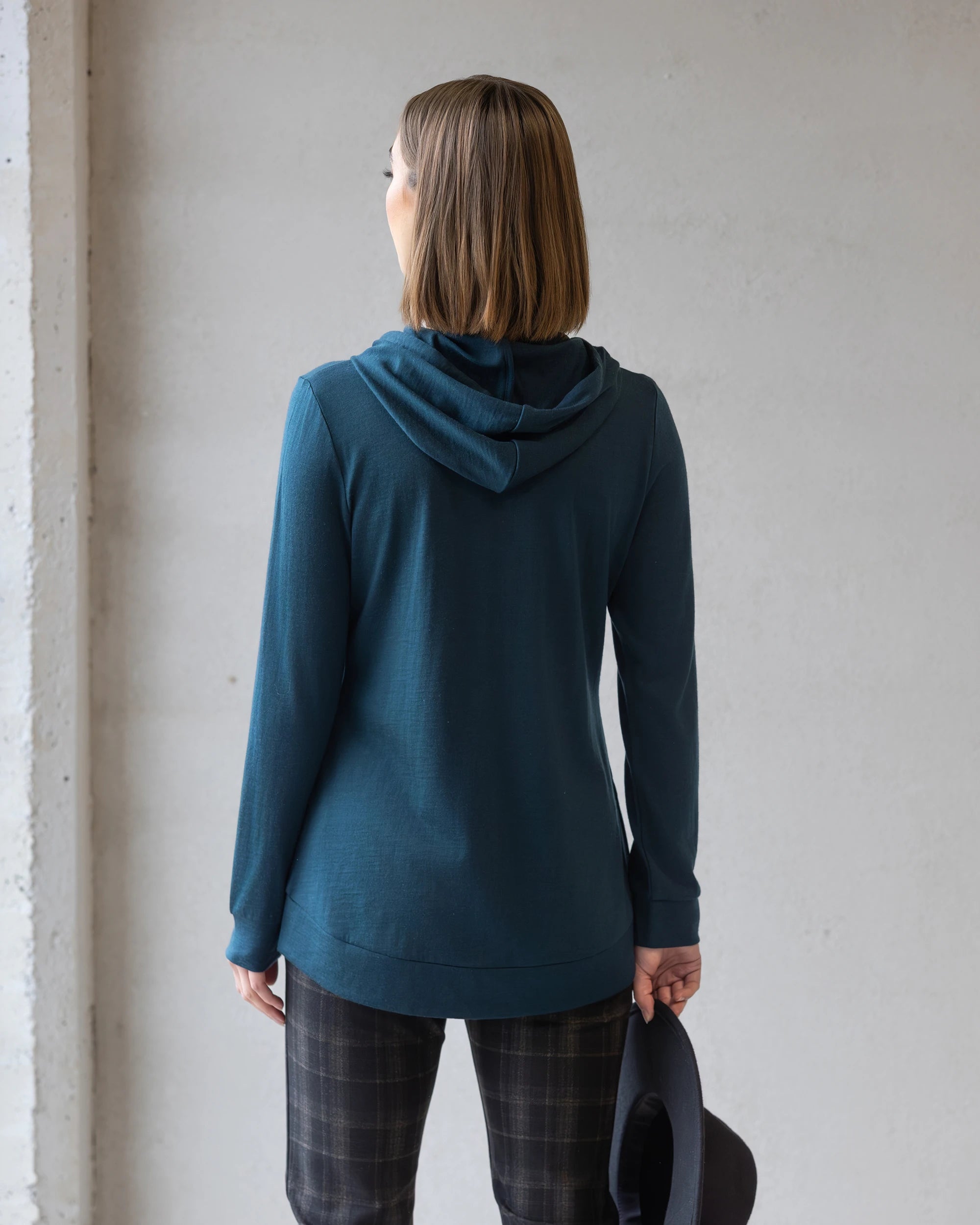 A person with shoulder-length brown hair, seen from behind, wears the Vassalli Shield Merino Hoodie in Dragonfly and plaid pants, holding a dark hat in their right hand against a plain light gray wall.