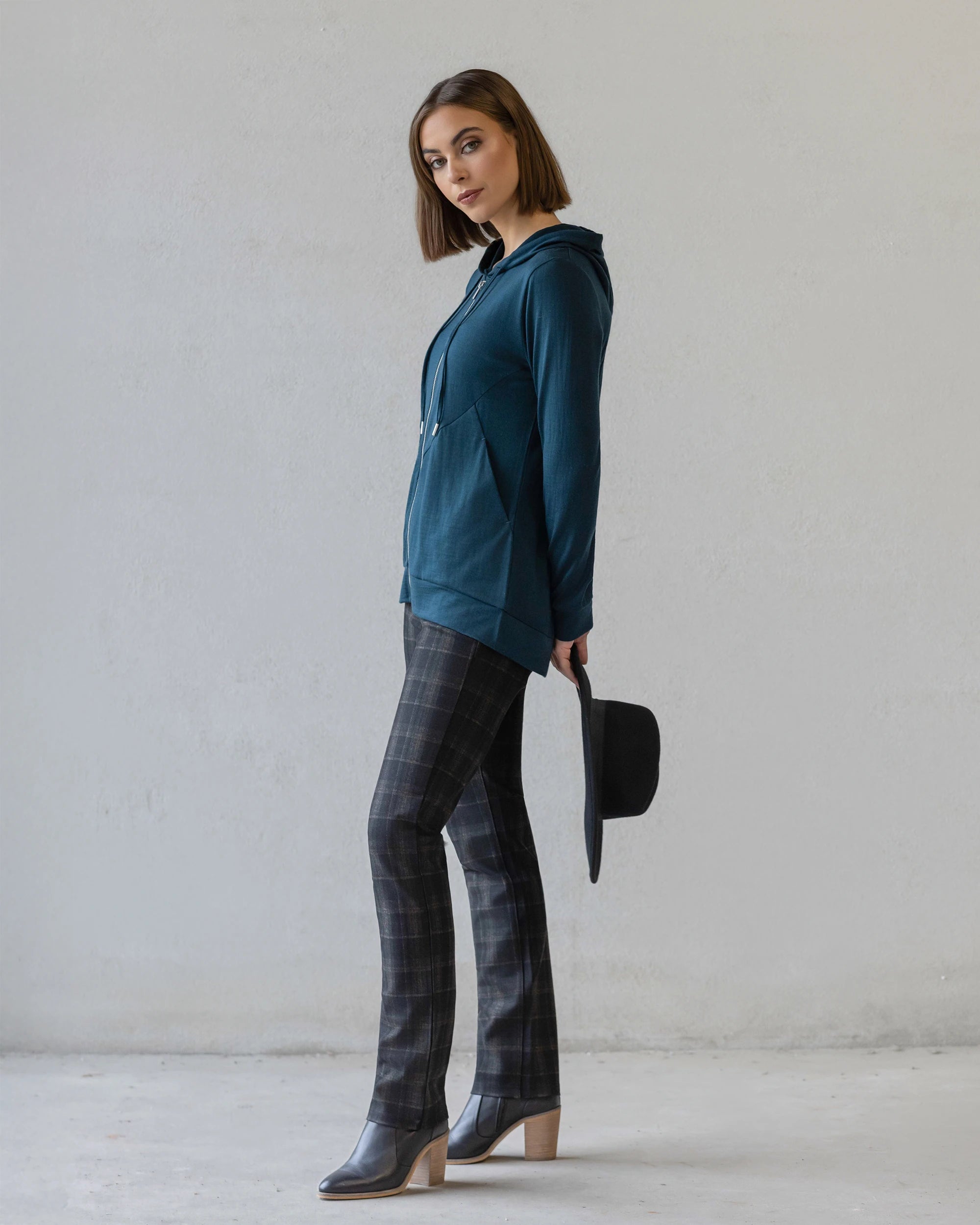 A woman stands sideways against a plain wall, wearing the Vassalli Shield Merino Hoodie in Dragonfly with plaid pants and heeled boots. She holds a black hat and faces the camera with a neutral expression.