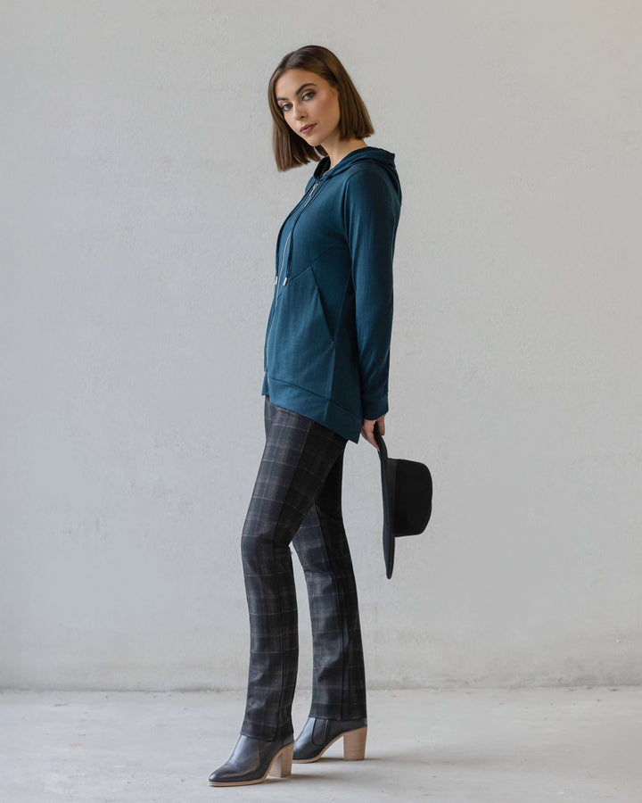 A woman stands sideways against a plain wall, wearing the Vassalli Shield Merino Hoodie in Dragonfly with plaid pants and heeled boots. She holds a black hat and faces the camera with a neutral expression.