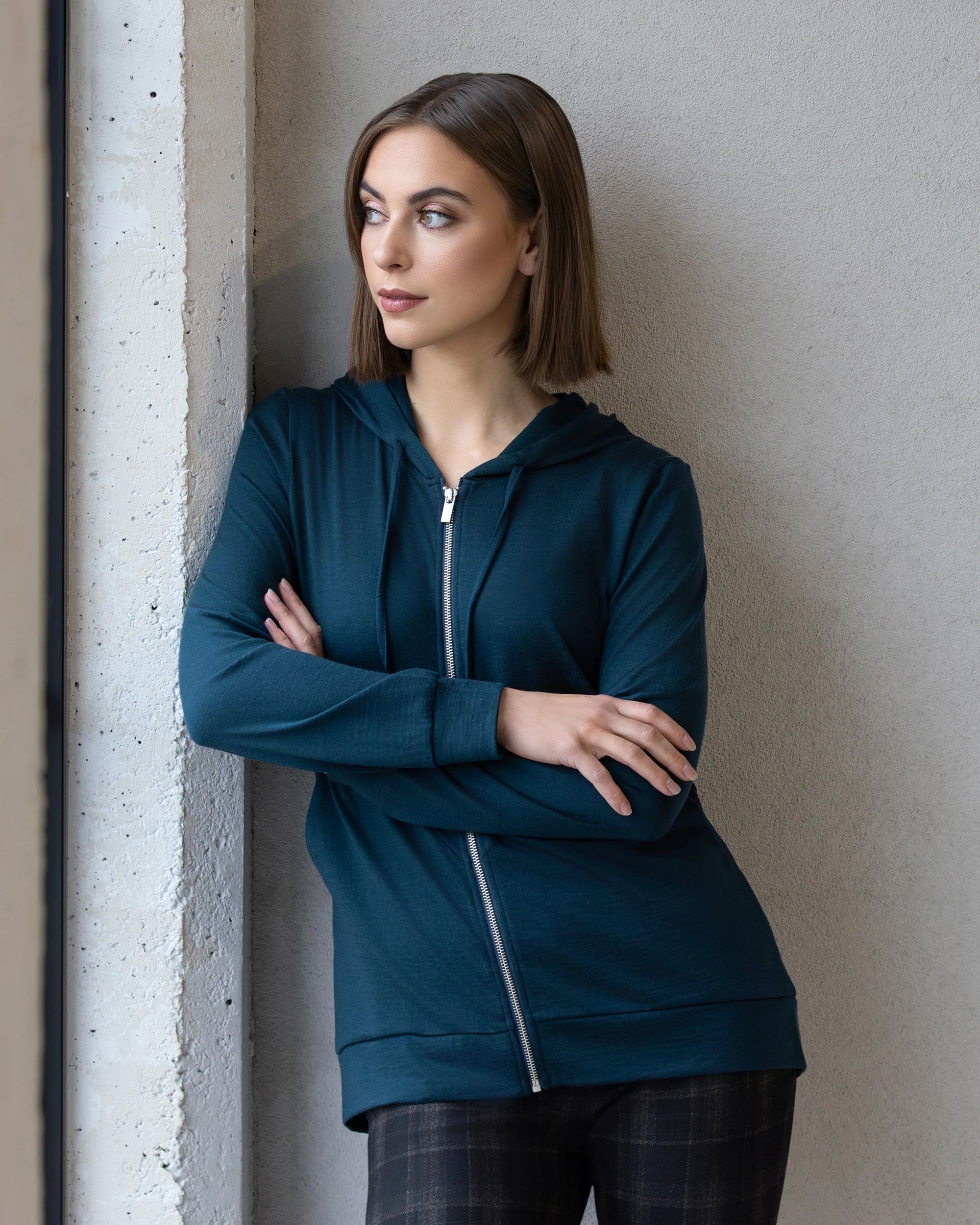 A woman with straight brown hair, wearing the Vassalli Shield Merino Hoodie in Dragonfly, stands with arms crossed and plaid pants, leaning against a light-colored wall and gazing thoughtfully out a nearby window.