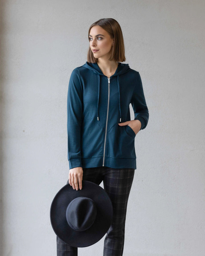 A woman in the Vassalli Shield Merino Hoodie - Dragonfly stands against a light gray wall, wearing plaid pants, holding a wide-brimmed black hat in one hand and looking off to the side.