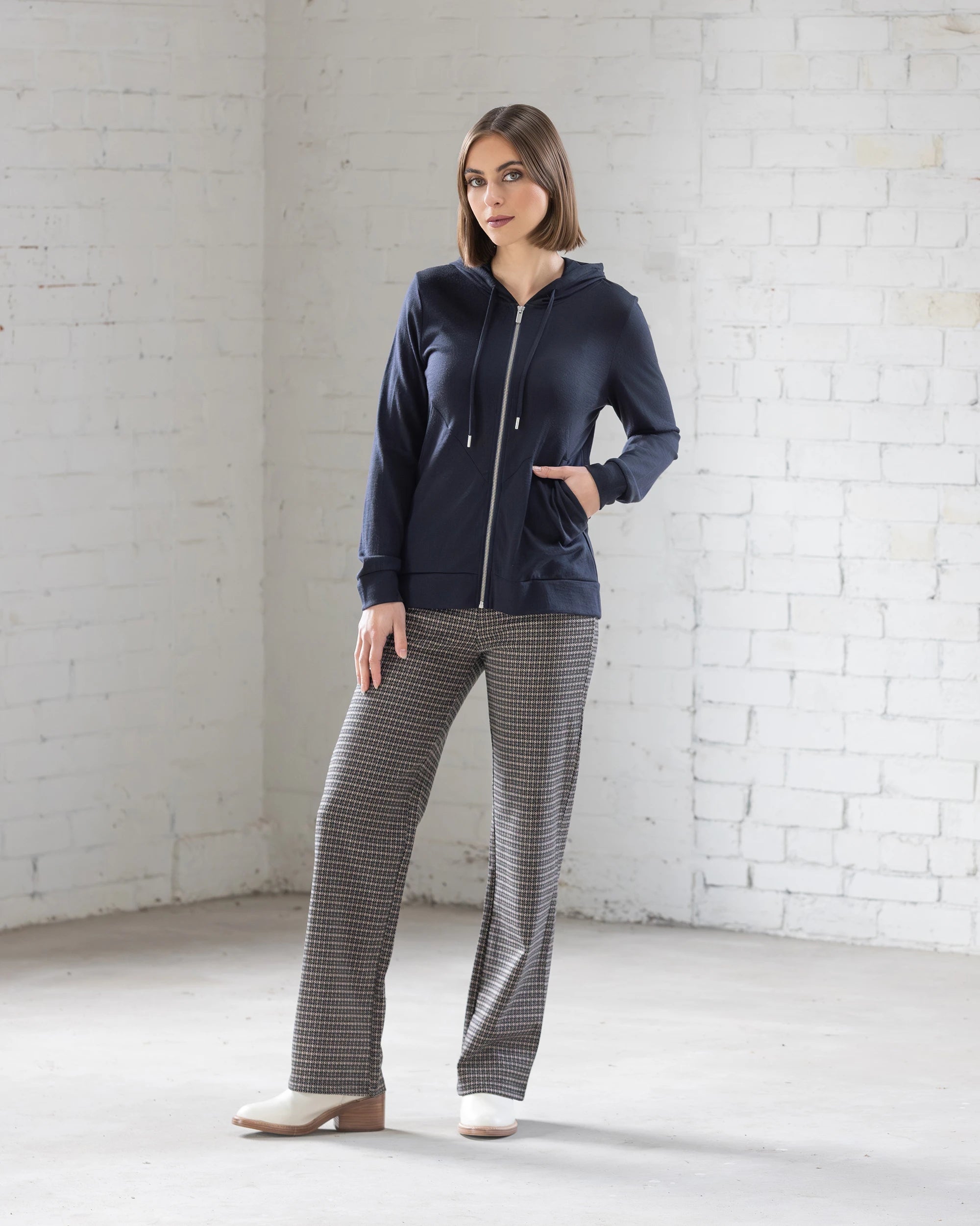 A woman stands indoors against a white brick wall, wearing the Vassalli Shield Merino Hoodie in Midnight with gray plaid pants and white shoes, one hand in her pocket and gazing slightly to the side.