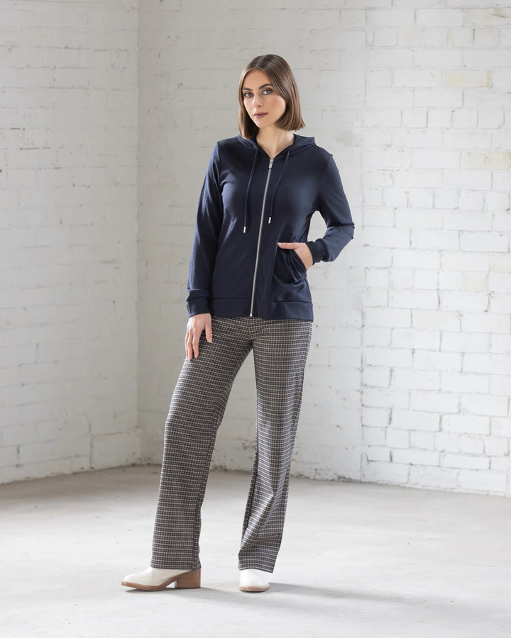 A woman stands indoors against a white brick wall, wearing the Vassalli Shield Merino Hoodie in Midnight with gray plaid pants and white shoes, one hand in her pocket and gazing slightly to the side.