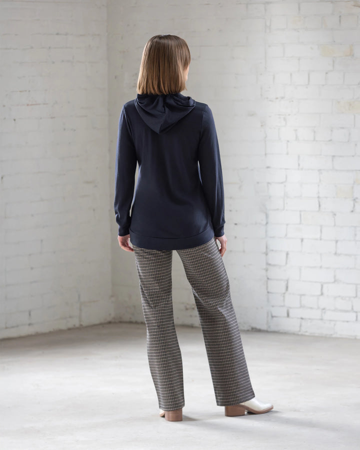 A person with straight, shoulder-length hair stands indoors facing a white brick wall, wearing the Vassalli Shield Merino Hoodie in Midnight, paired with plaid pants and white shoes.