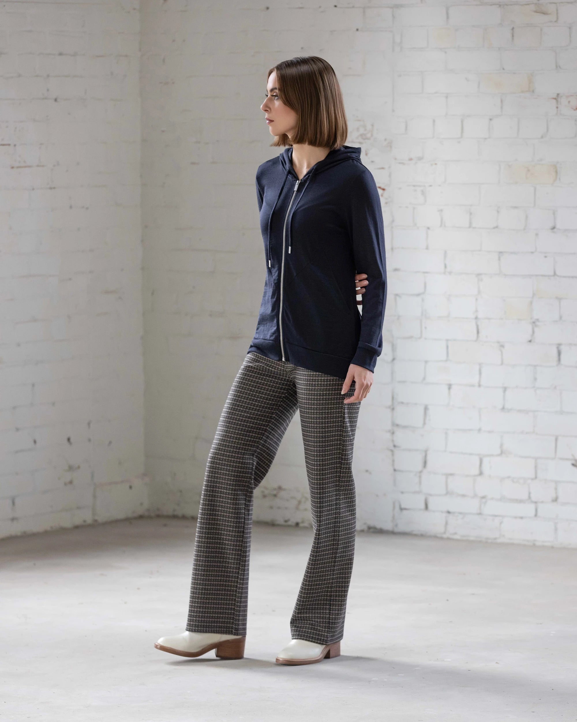 A woman stands indoors against a white brick wall, wearing the Vassalli Shield Merino Hoodie in Midnight, paired with checkered trousers and white heels. She looks to the side with her hands behind her back.