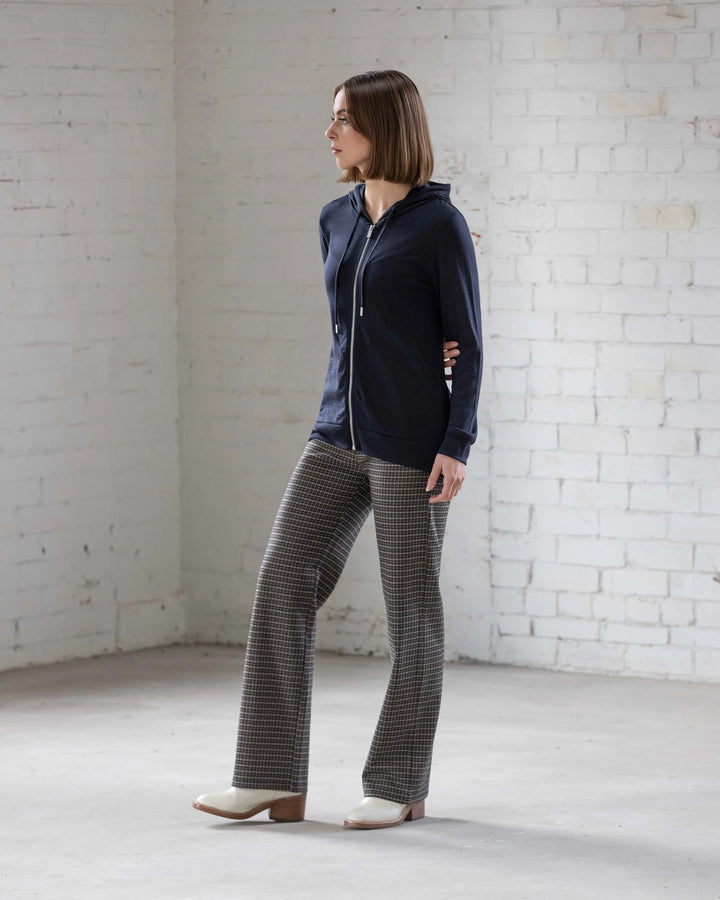 A woman stands indoors against a white brick wall, wearing the Vassalli Shield Merino Hoodie in Midnight, paired with checkered trousers and white heels. She looks to the side with her hands behind her back.