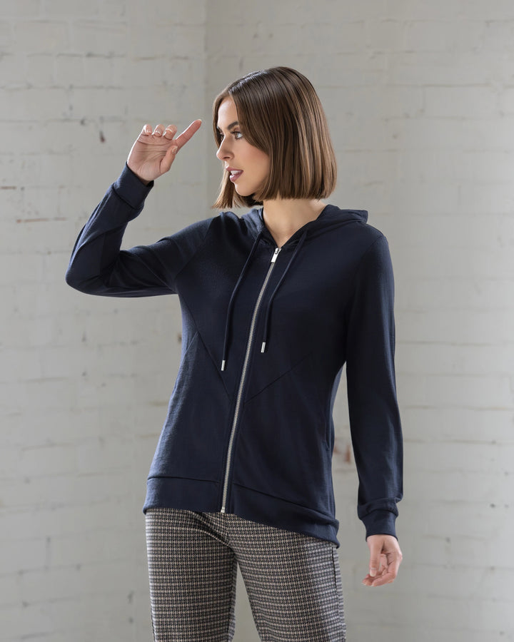 A woman wearing the Vassalli Shield Merino Hoodie in Midnight stands by a white brick wall, looking to the side with her left hand near her forehead.