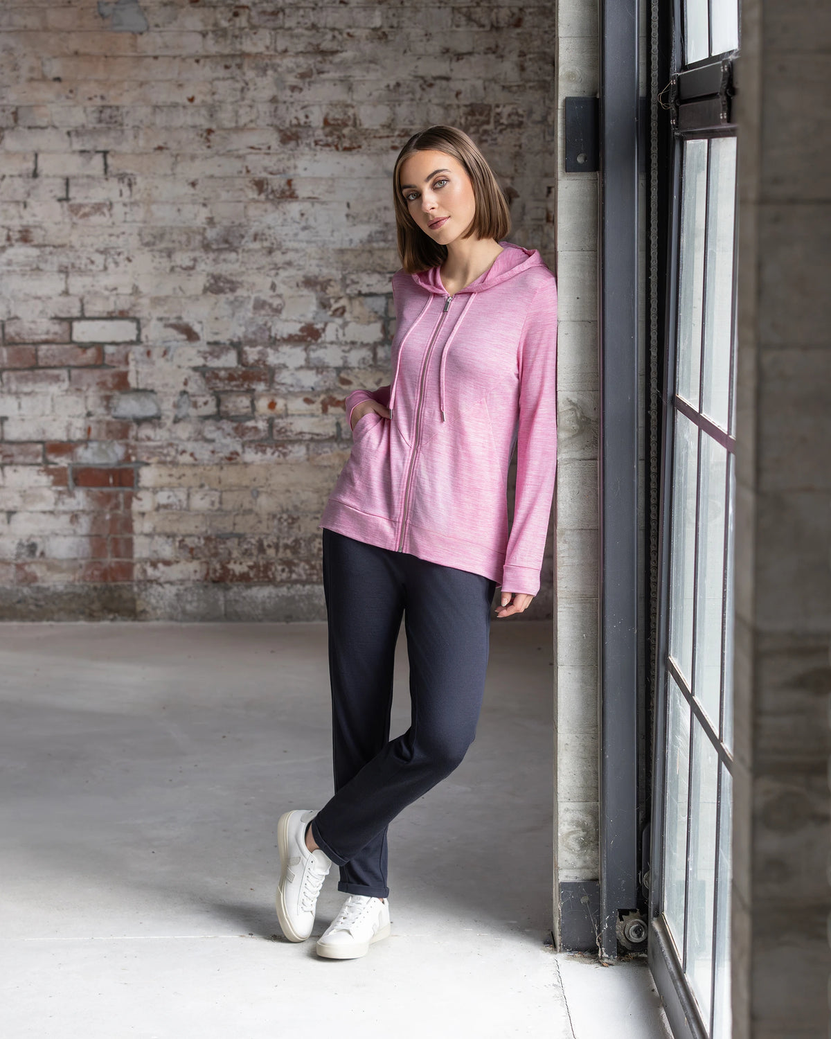 A woman in the Vassalli Shield Merino Hoodie in Pink Marle, black pants, and white sneakers leans against a large window in an industrial-style room—ideal for winter layering.