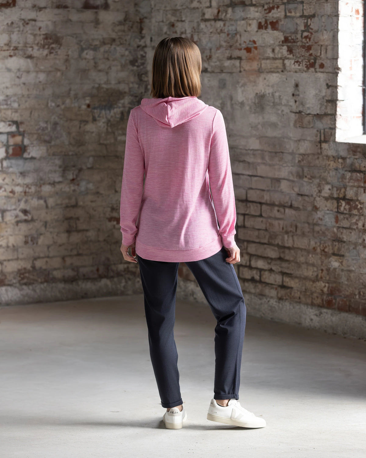 A person with shoulder-length brown hair wears the Vassalli Shield Merino Hoodie in Pink Marle, paired with dark pants and white sneakers, standing indoors by a distressed brick wall with a window—ideal for cozy winter layering.