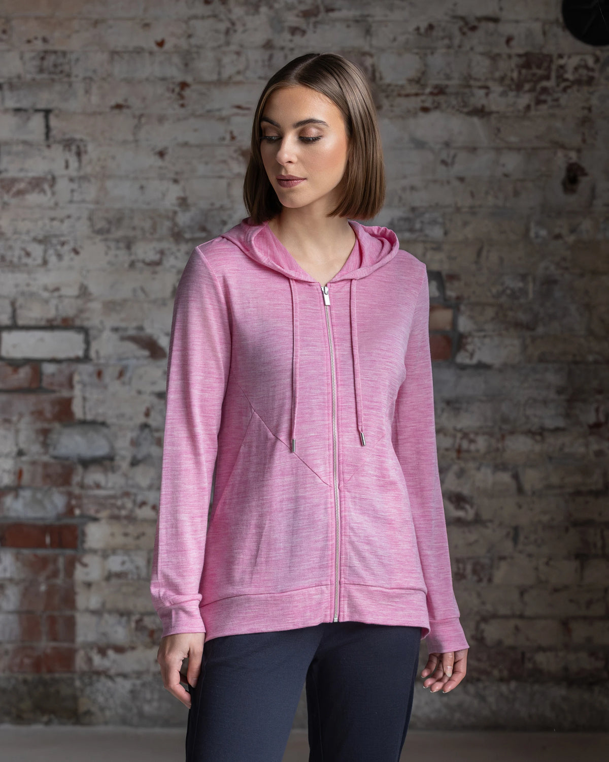 A woman with straight brown hair wears the Vassalli Shield Merino Hoodie in Pink Marle and dark pants, standing before a textured brick wall, looking down with a neutral expression—ideal for winter layering.