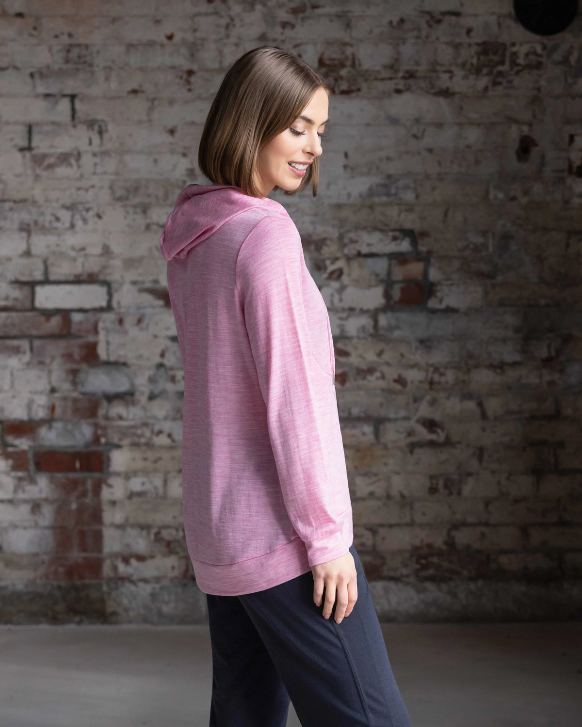 A woman wears the Vassalli Shield Merino Hoodie in Pink Marle with dark pants, standing sideways and smiling softly against a brick wall—ideal for cozy winter layering.