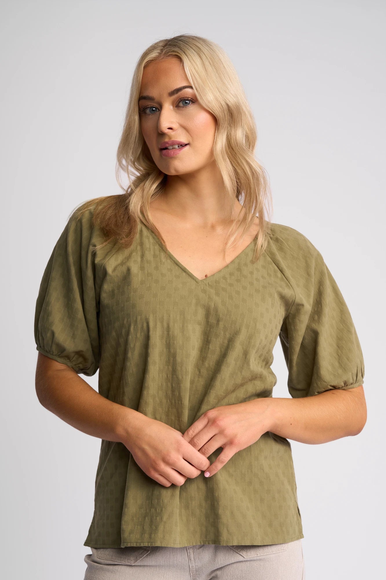 A woman with long blonde hair wears a Vassalli Pitch Top in Khaki featuring short puff sleeves and a V-neck, standing against a plain light gray background.